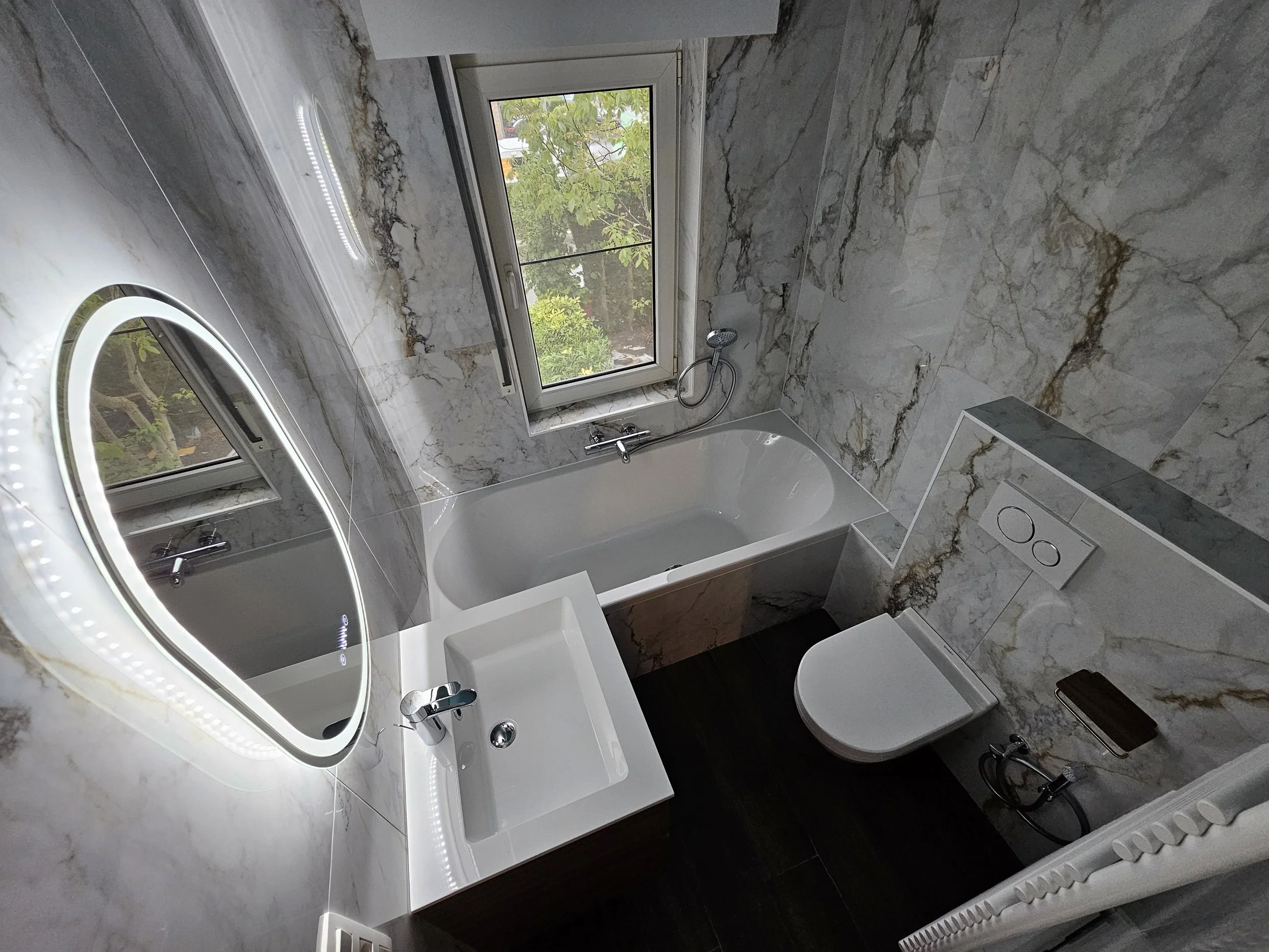 Moderne badkamer met muur- en vloertegels met opvallende marmerprint, een wit bad, een klein wit wastafelmeubel met spiegel, een toilet met ingebouwde spoelknop, een open raam, en een handdouche naast het bad onder het raam.