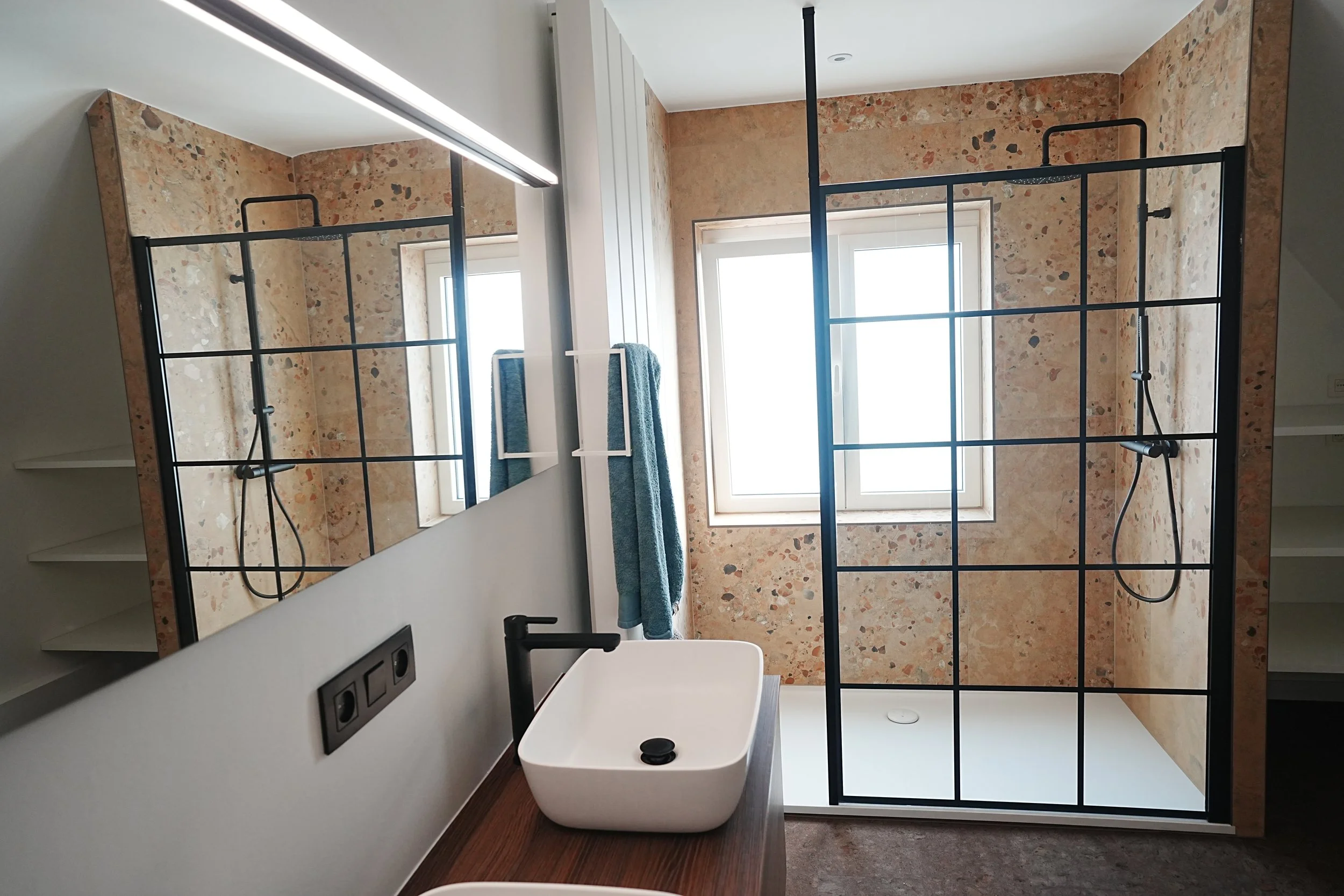 Badkamer met granieten muur, twee ramen, een zwart metalen inloopdouche met glas, een witte wasbak met zwarte kraan, een handdoekrek met blauwe handdoek en spiegel