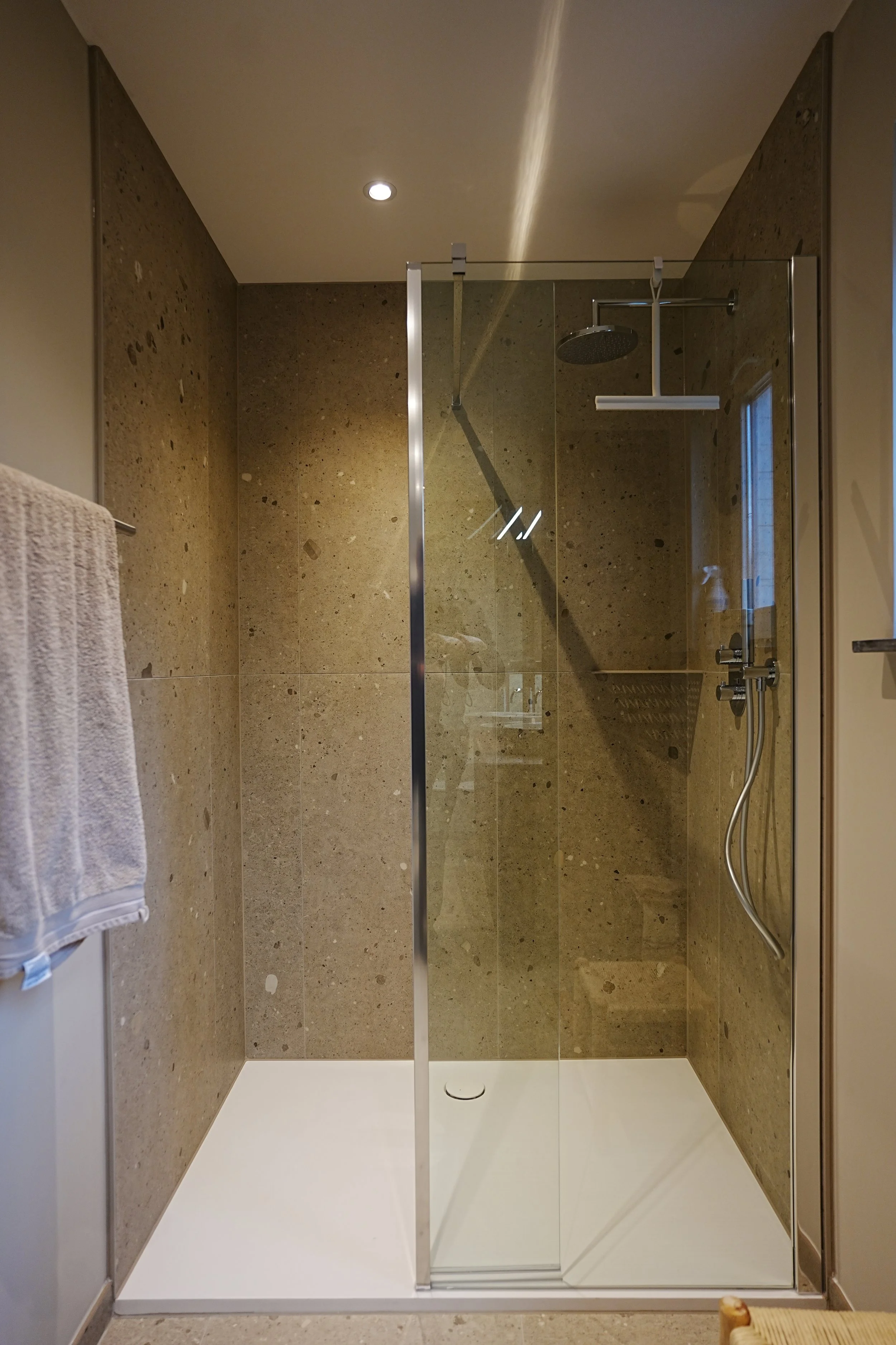 Een inloopdouche met glazen wand en een regen-douchekop op een muur met beige marmerachtige tegels. Rechts een thermostatische douchekraan met een flexibele douchekop. Links een handdoek aan de muur.