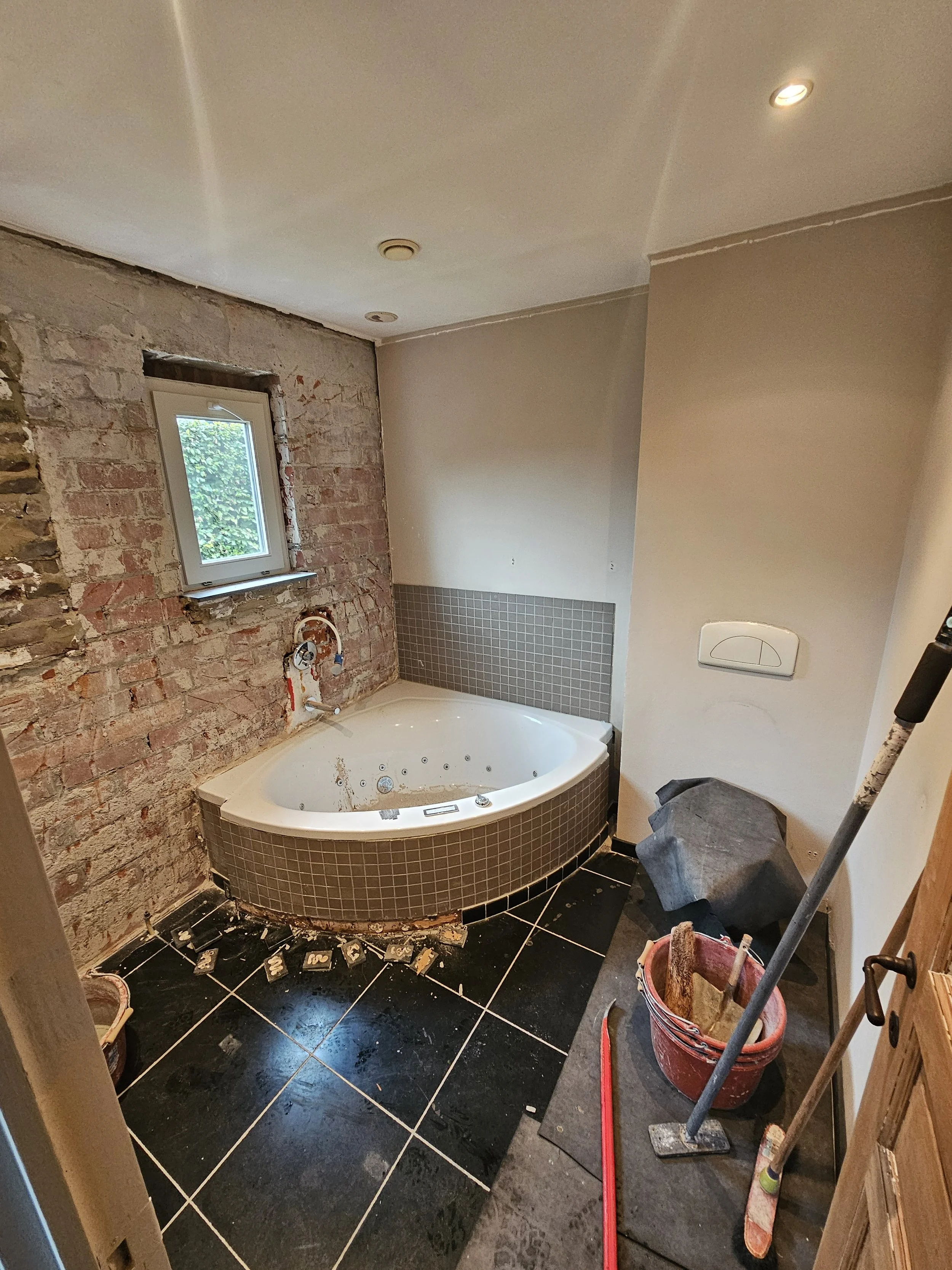 Een badkamer in aanbouw met een ingebouwde hoekbadkuip, een klein raam, en bouwmaterialen en gereedschap op de vloer.