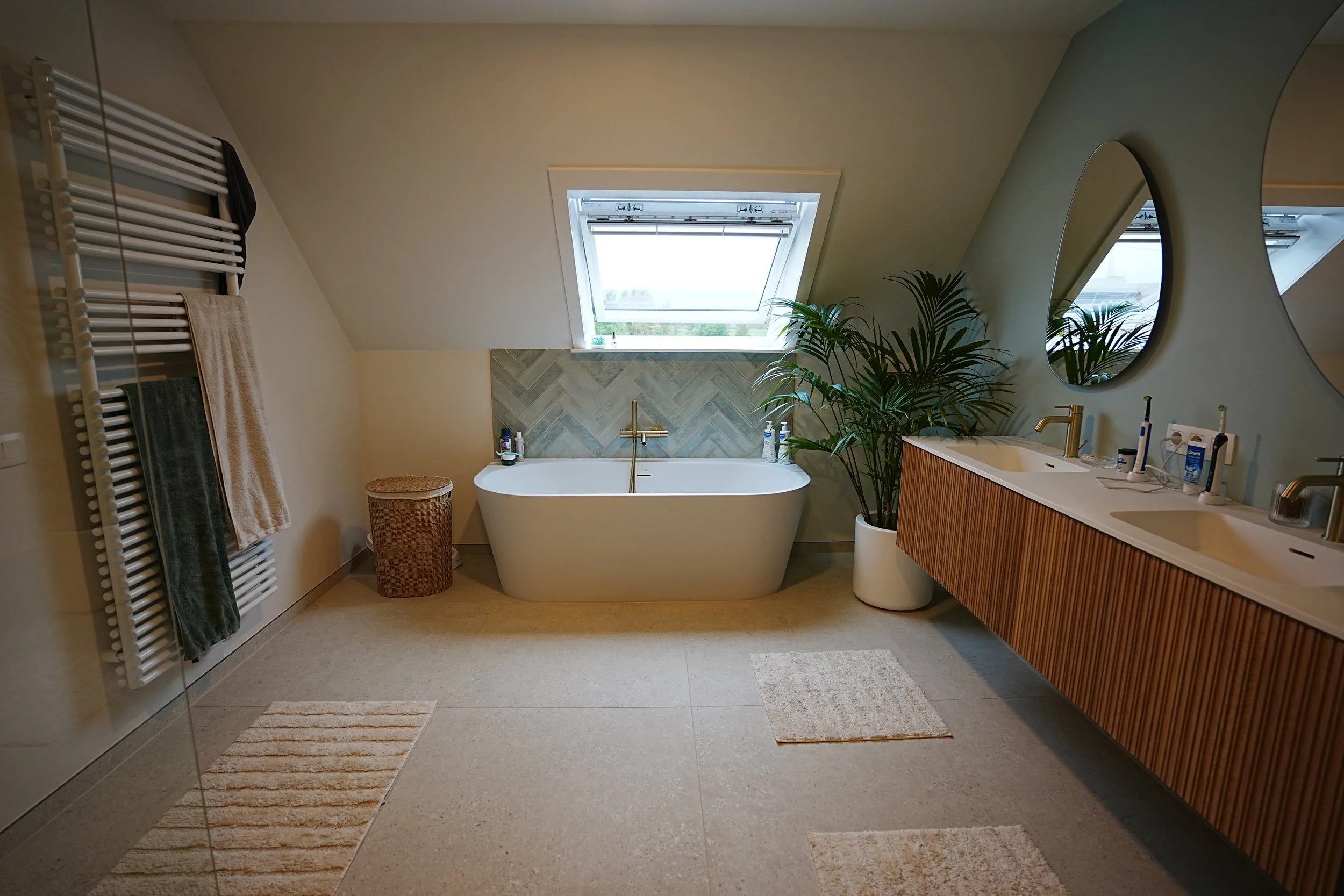 Moderne badkamer met vrijstaand bad onder een schuine dakvenster, grote spiegel boven dubbele wastafel, planten, handdoekradiator en neutrale kleuren.
