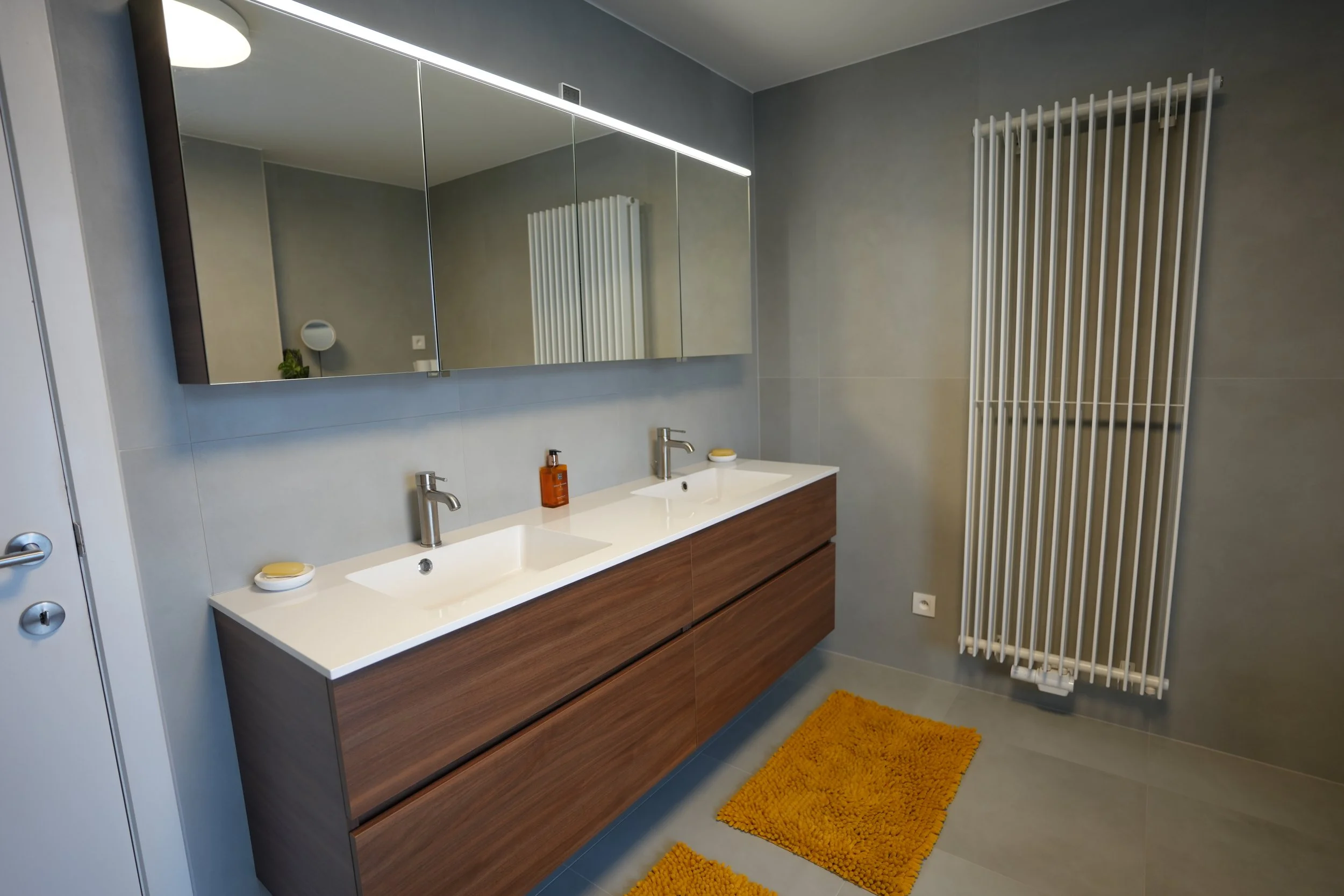 Moderne badkamer met dubbele wastafel, grote spiegelkast, radiator en gele badmatten.