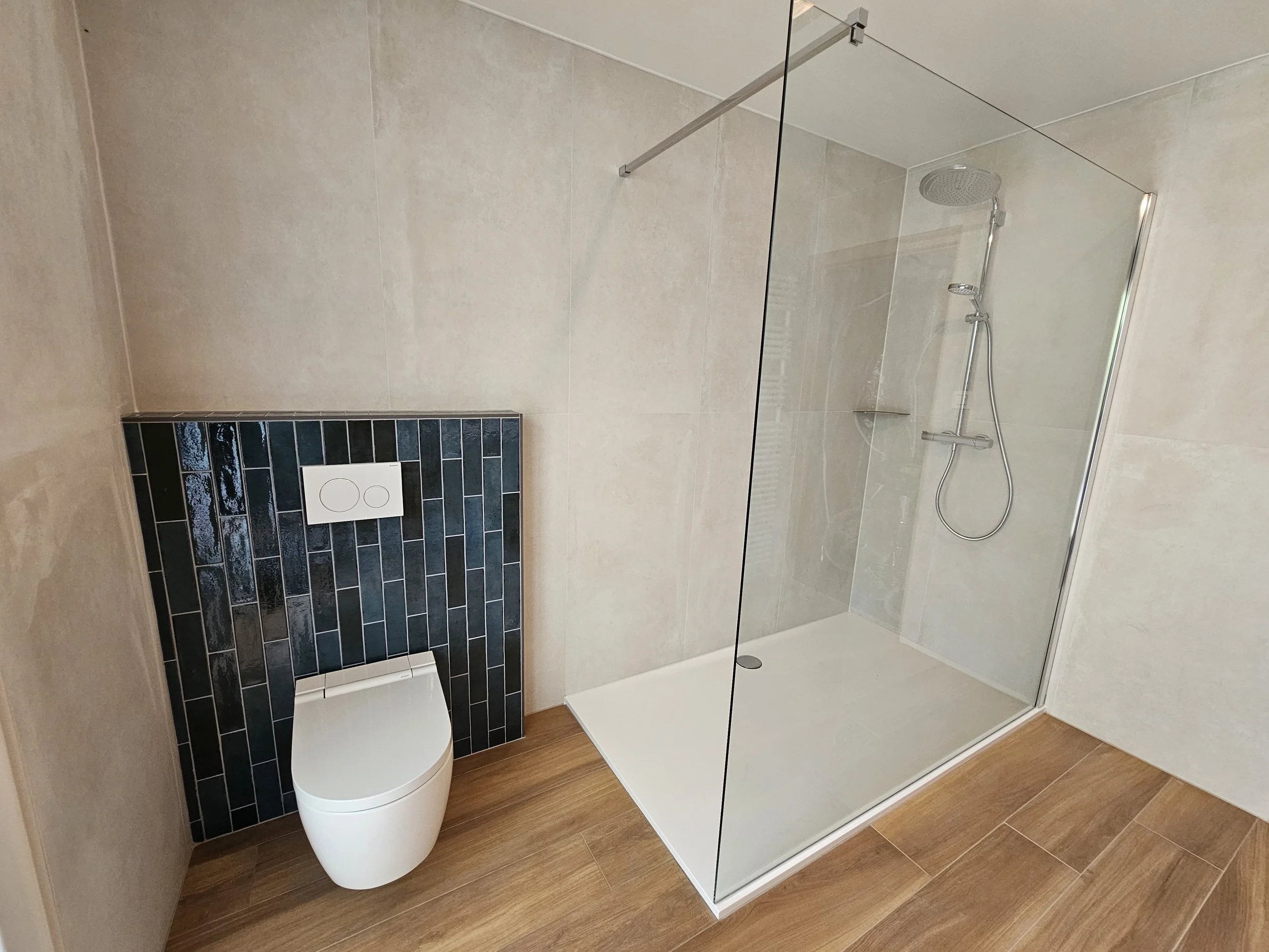 Moderne badkamer met toilet en inloopdouche, met lichte muren, houten vloer en donkerblauwe tegels achter het toilet.