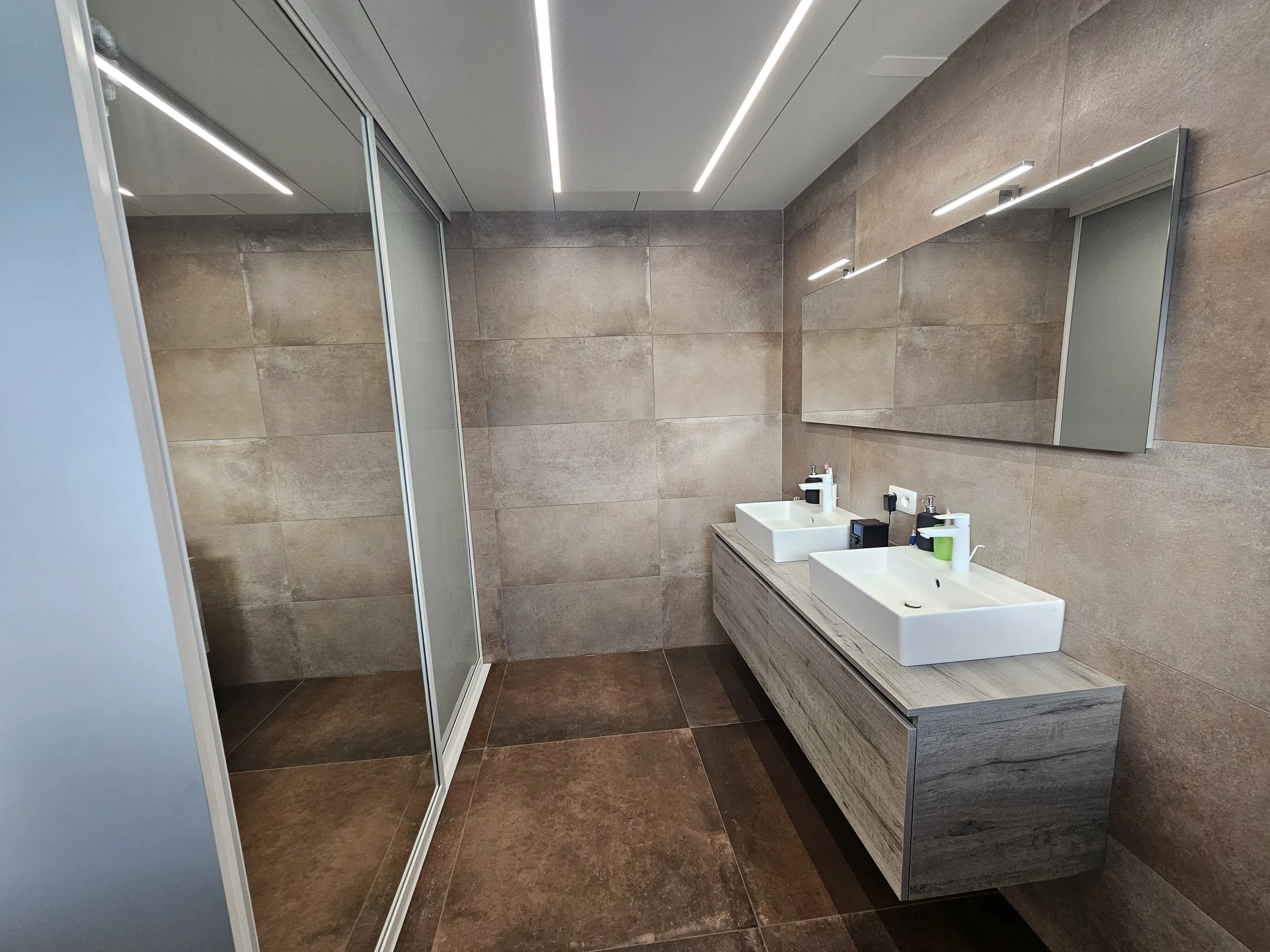 Moderne badkamer met dubbele wastafels, grote spiegel, beige tegelwanden en -vloeren, en een inloopdouche met glasdeuren.