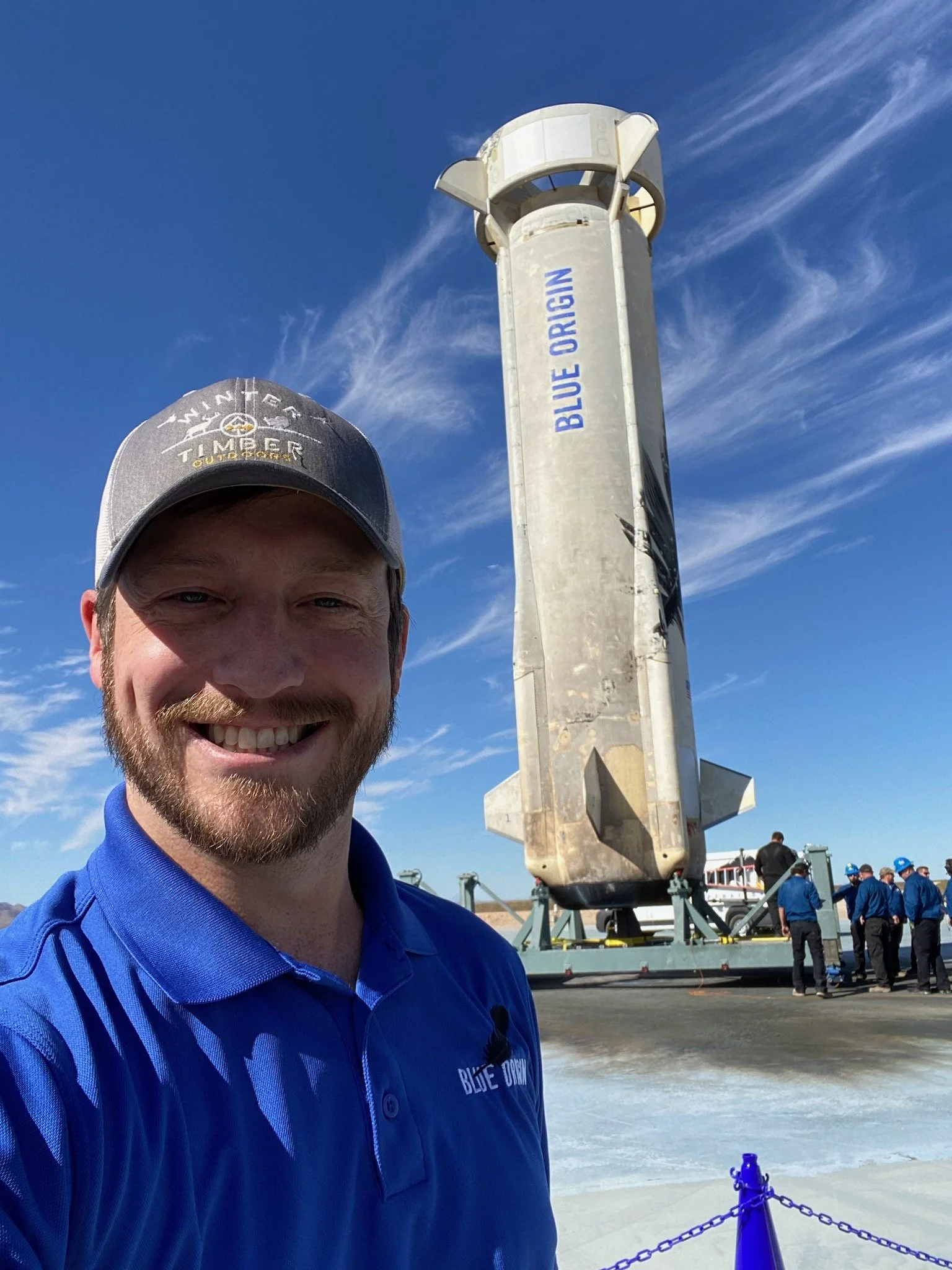 A man in a blue shirt and cap taking a selfie in front of a SpaceX Starship prototype on a launch pad, with a clear blue sky and wispy clouds.