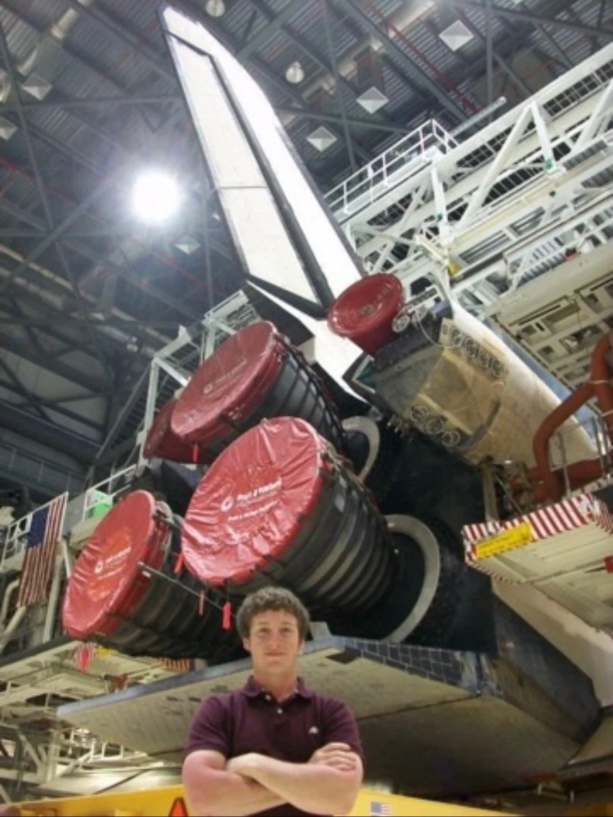A person standing in front of a spacecraft or space vehicle inside a large industrial or research facility. The spacecraft has several large red and black components covered with red protective caps, and is surrounded by metallic structures and scaffolding. The person's arms are crossed, and they wear a dark purple polo shirt. The ceiling is high with visible metal beams and skylights.