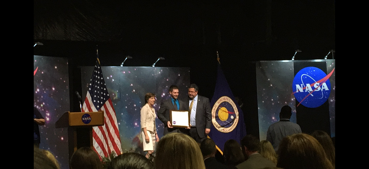 Travis Davis standing on a stage at NASA award ceremony, with NASA logos, flags, and a starry background.