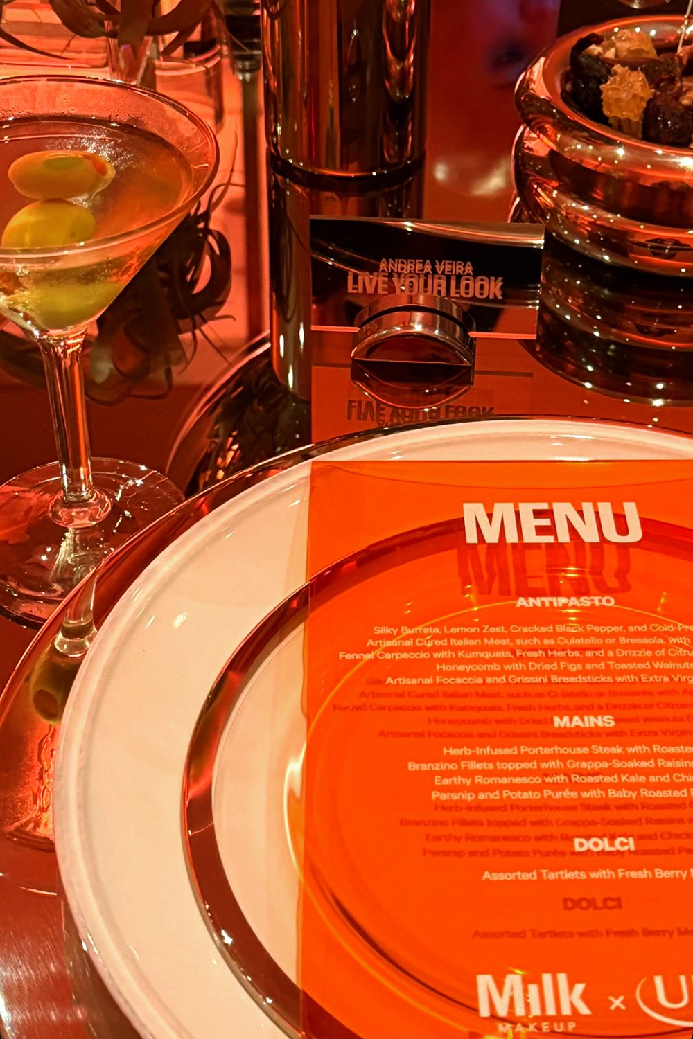 Close-up of a guest's place setting at the Milk Makeup x Ulta Beauty dinner, featuring an orange acrylic menu by Cava & Cake with Italian-inspired dishes, next to a martini glass and a custom “Live Your Look” name card.