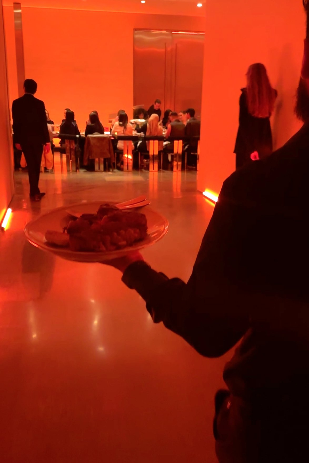 Server carrying a platter of food through a glowing orange-lit hallway toward a long dinner table filled with guests at the Milk Makeup x Ulta Beauty dinner party at Milk Studios in Los Angeles.