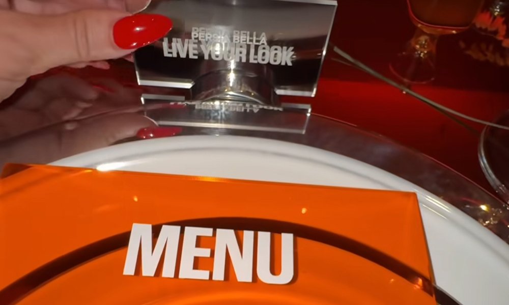 Close-up of a glam place setting at the Milk Makeup x Ulta Beauty dinner, featuring a bold orange menu, a name card that reads “LIVE YOUR LOOK,” and perfectly manicured red nails holding the acrylic card at Milk Studios, Los Angeles.
