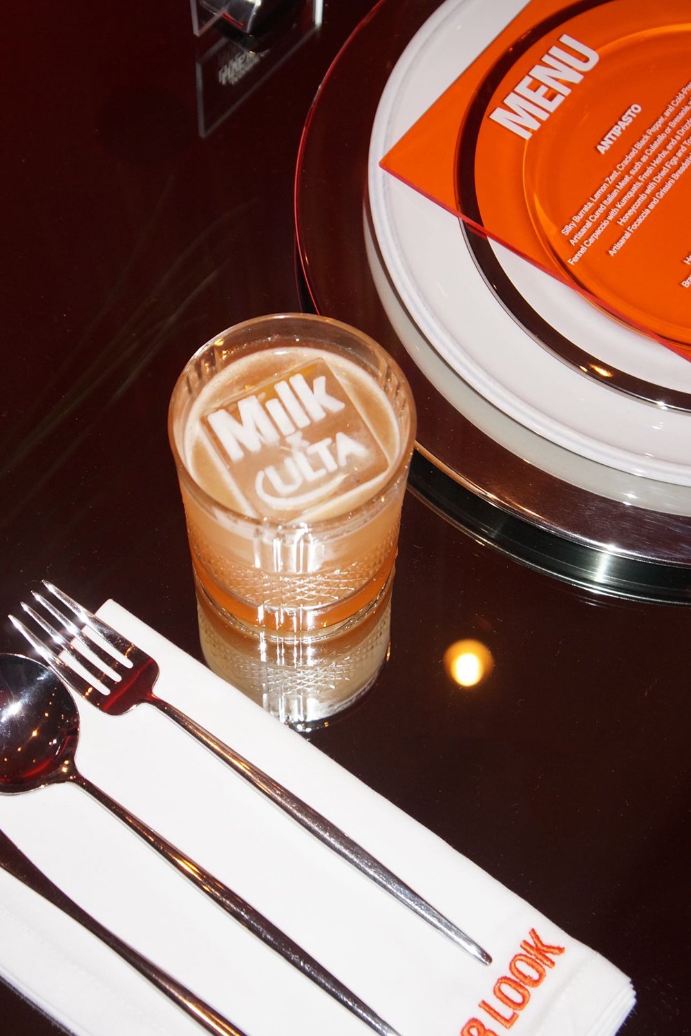 Close-up of a branded cocktail with custom ice featuring the Milk Makeup and Ulta logos, set beside a modern place setting and bold orange event menu at the Milk Studios dinner.