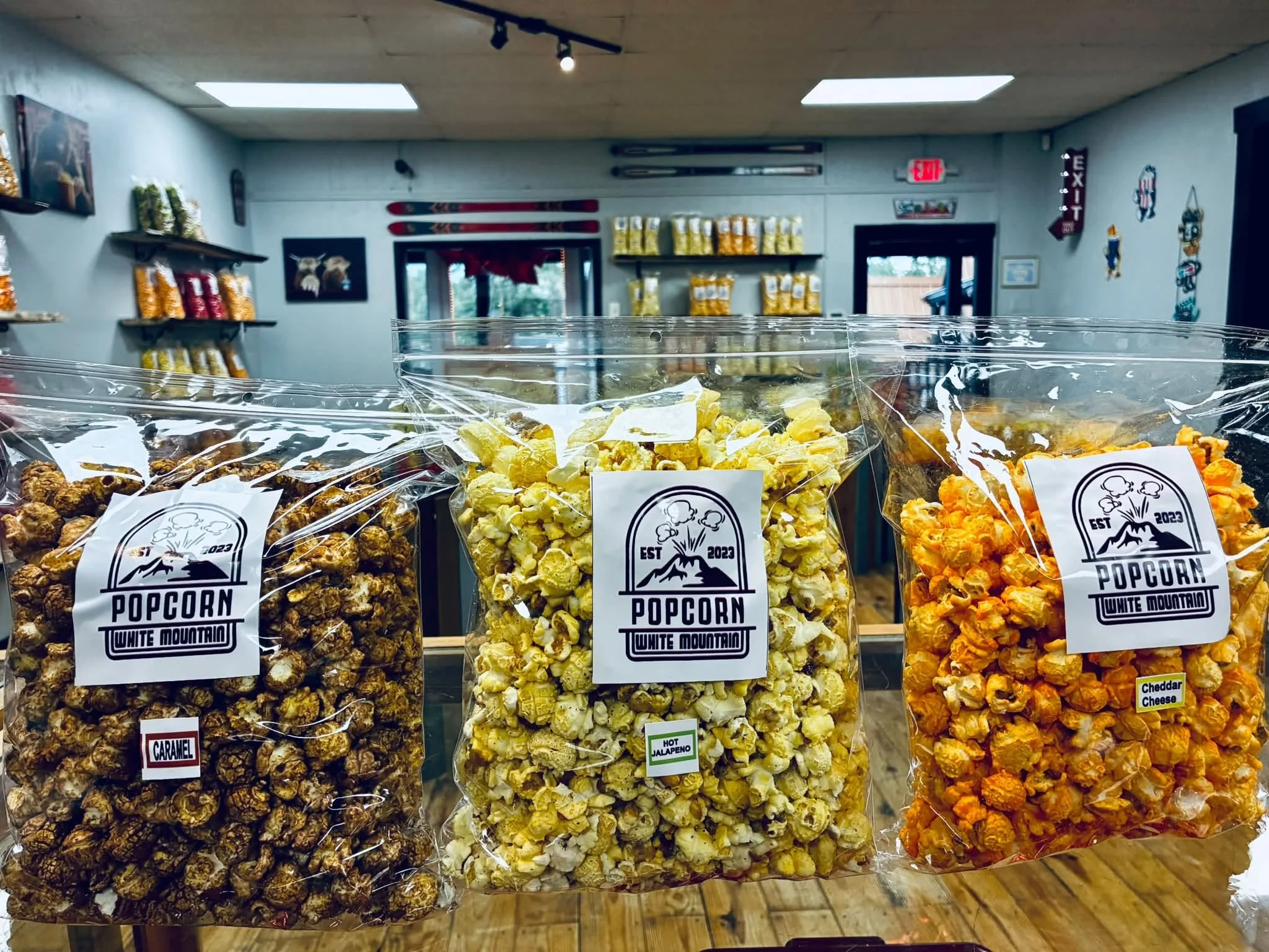 Three bags of White Mountain Popcorn’s gourmet popcorn—caramel, hot jalapeño, and cheddar cheese—displayed inside the Ruidoso shop with shelves of popcorn flavors in the background.