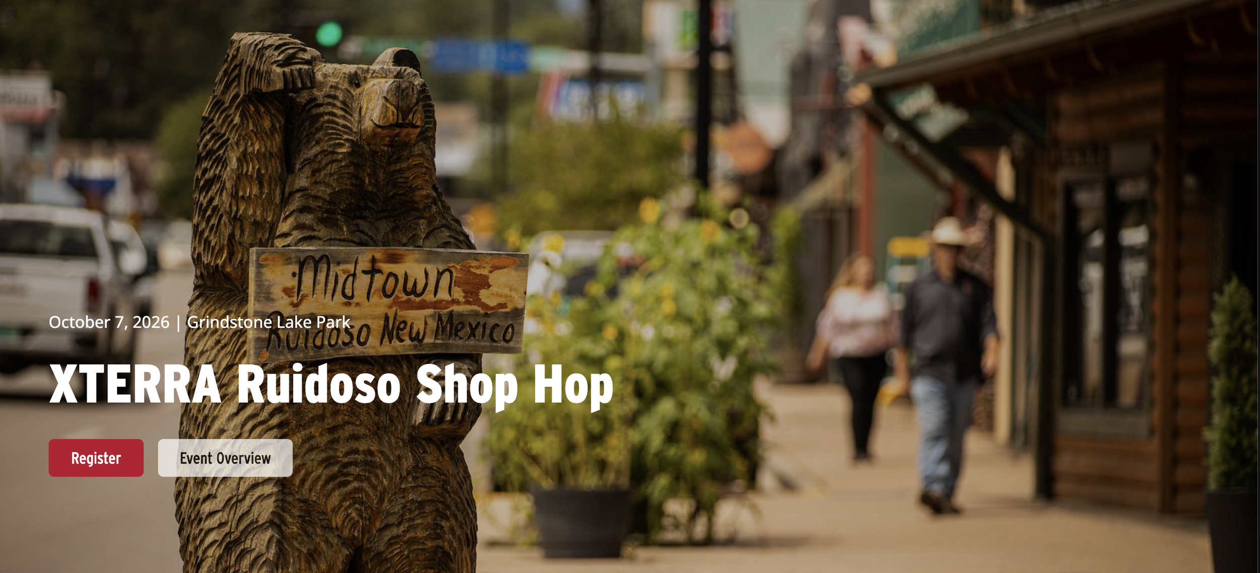 Wooden bear sculpture holding a “Midtown Ruidoso, New Mexico” sign along Sudderth Drive, with Midtown storefronts and pedestrians in the background during XTERRA World Championship week.