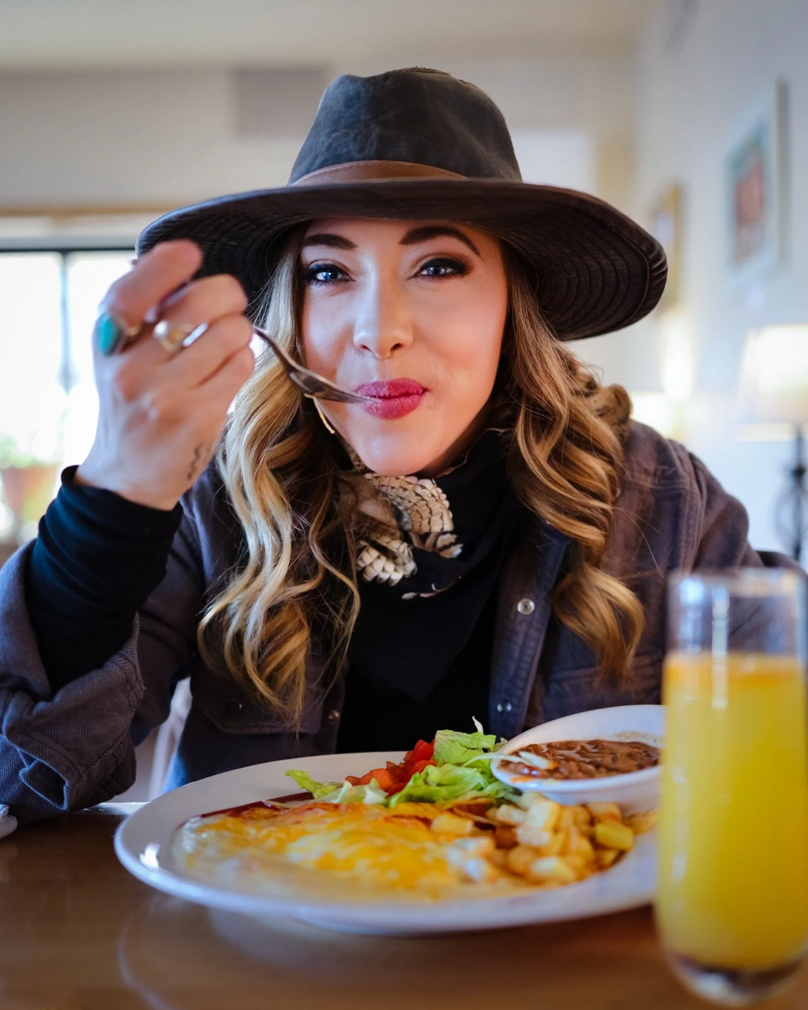 A woman in a wide-brimmed hat enjoys a bite of her meal at a restaurant, with a plate of New Mexican cuisine and a glass of orange juice in front of her.