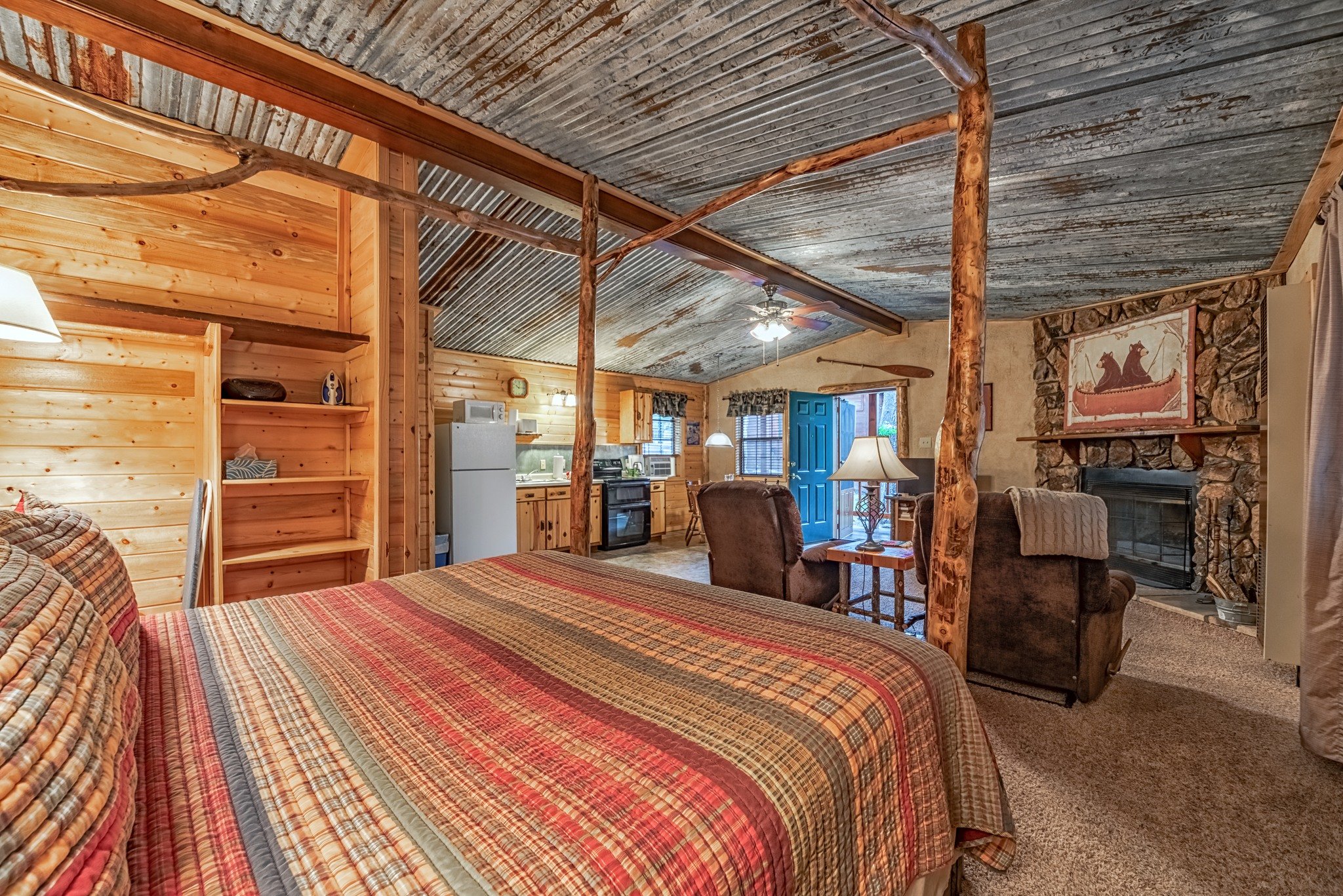 Cozy rustic cabin interior with wood-paneled walls, a metal corrugated ceiling, a stone fireplace, and an open layout featuring a bed, recliners, and a small kitchen.