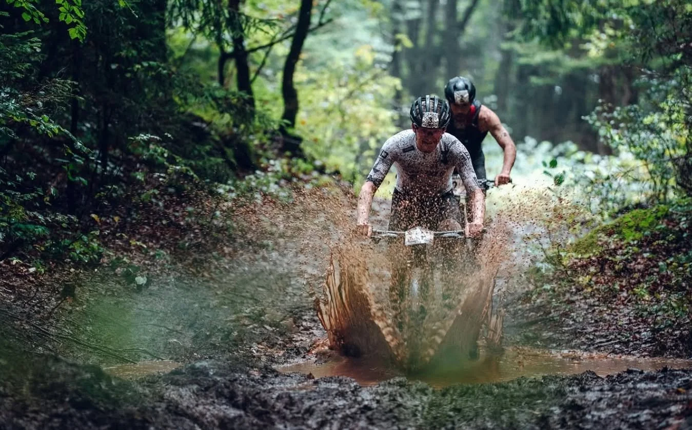 Mountain bikers race through a muddy forest trail during an XTERRA off-road cycling event.