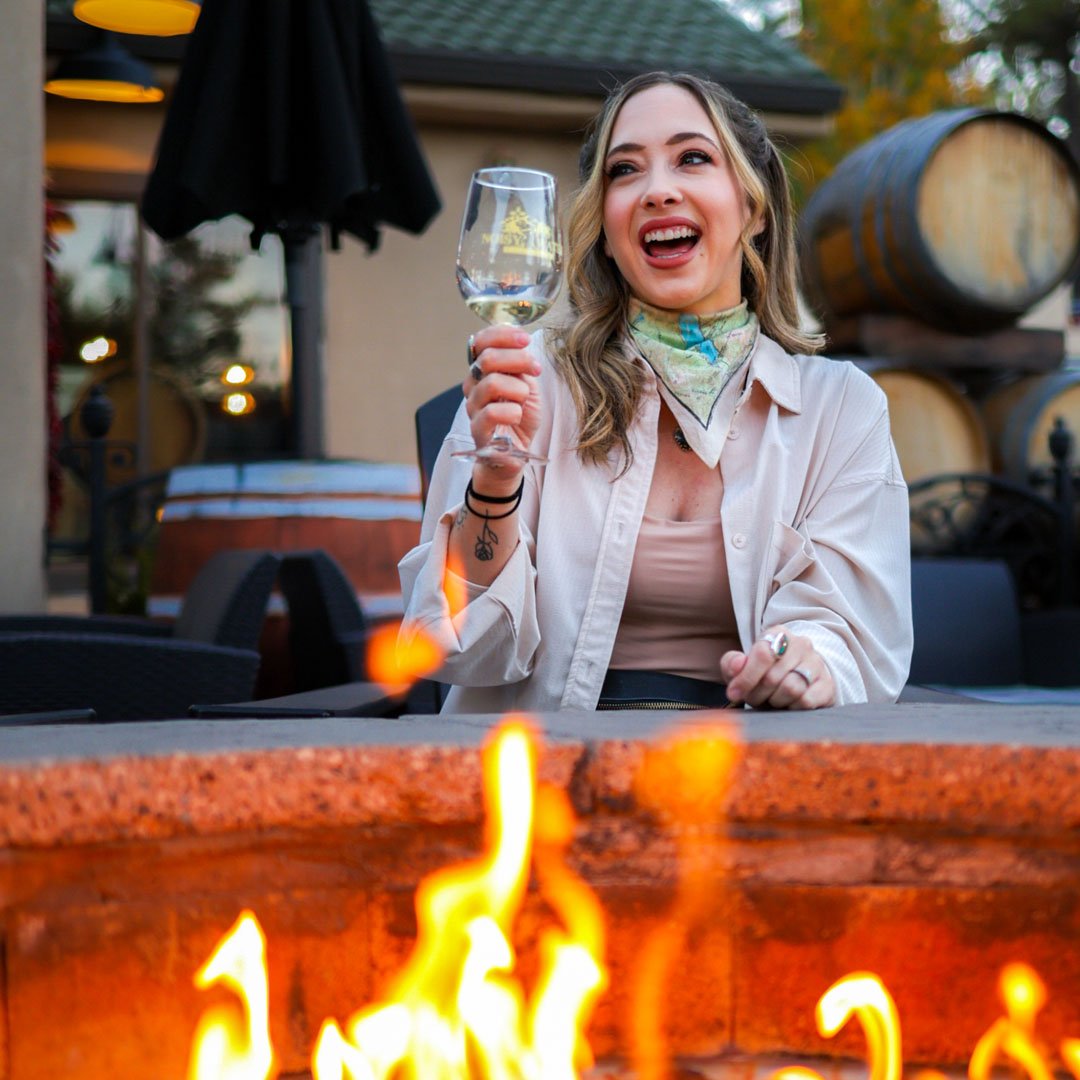 A visitor enjoys a glass of local wine beside a cozy outdoor fire pit at a Ruidoso winery. Surrounded by wine barrels and mountain scenery, the patio offers a relaxing place to unwind during a fall getaway in the Sacramento Mountains.