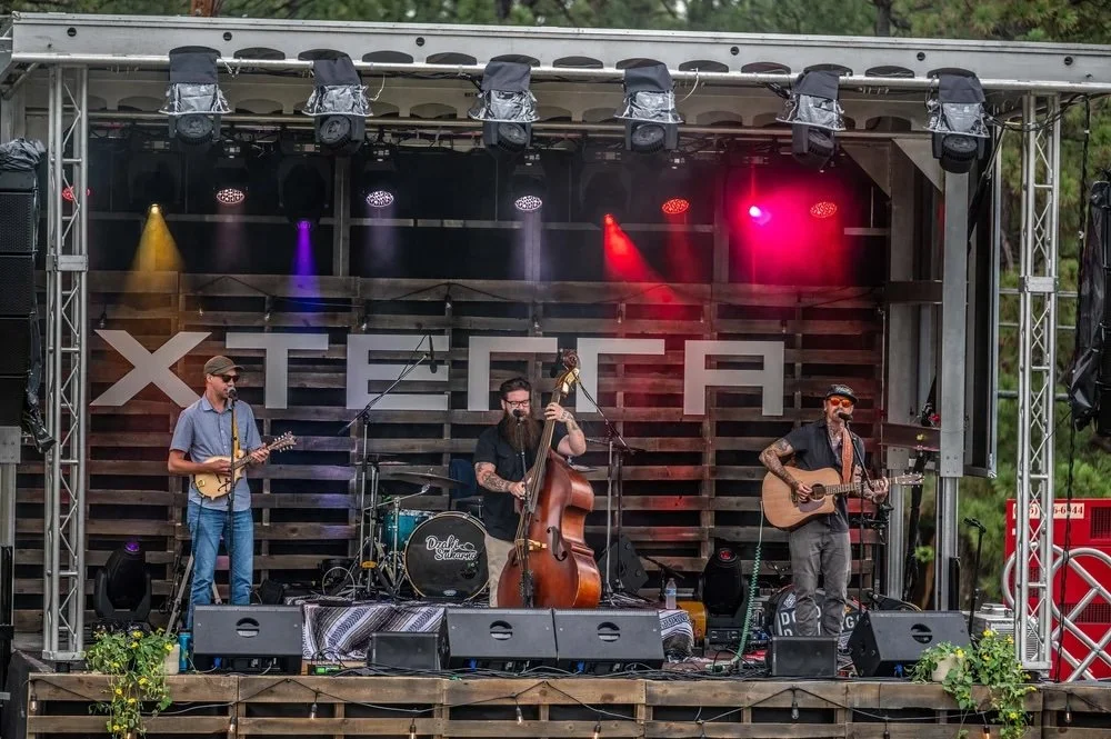 The Doso Dirtbags perform live on the XTERRA stage in Ruidoso, entertaining festivalgoers with bluegrass and Americana music during an outdoor mountain event surrounded by towering pine trees in the Sacramento Mountains.