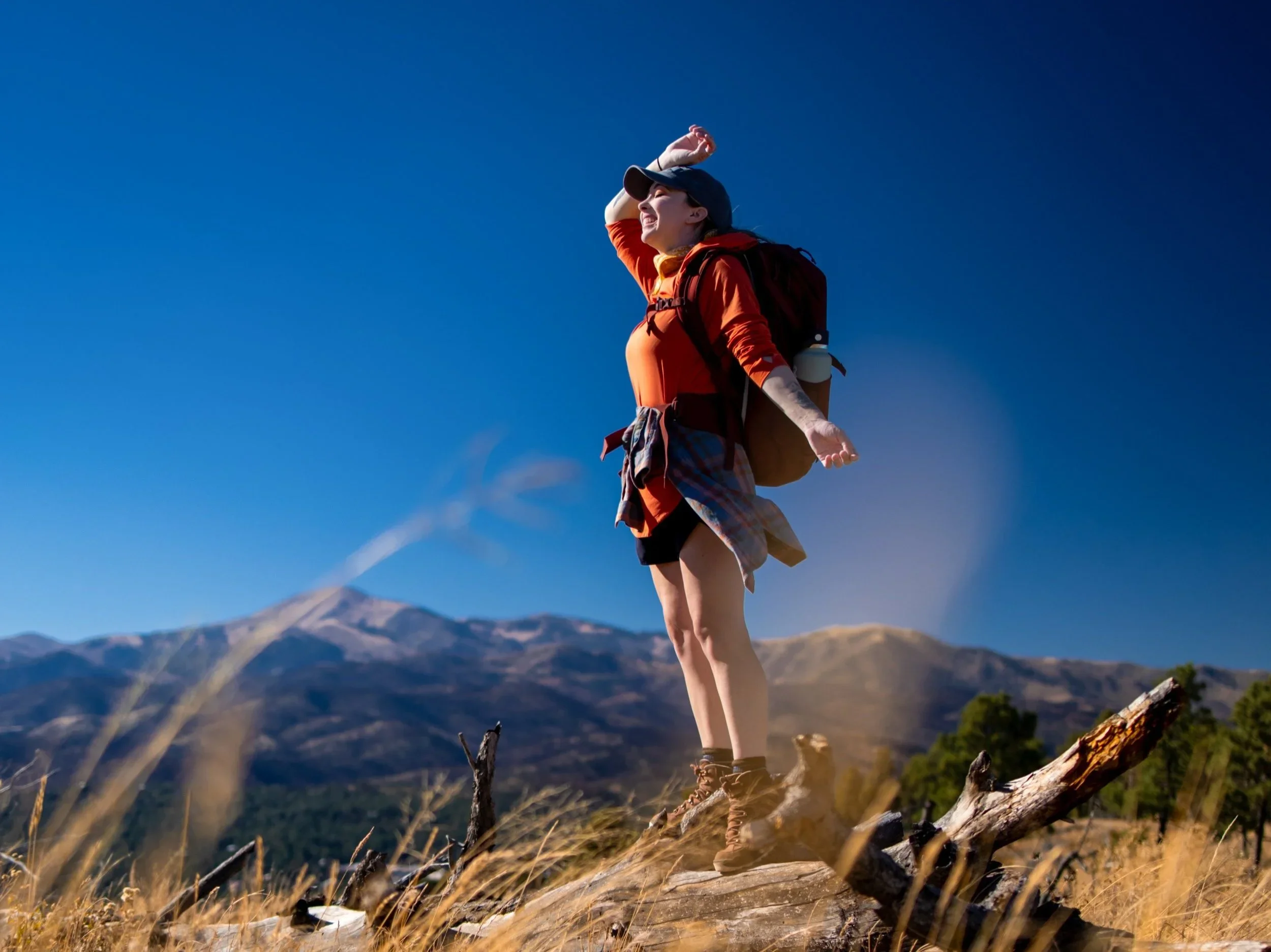 A hiker stands on a sunlit ridge with a backpack, taking in wide mountain views under a clear blue sky.