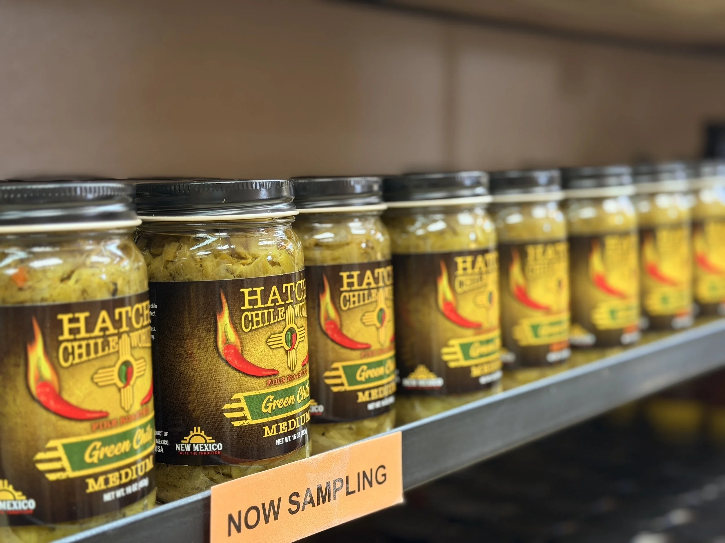 Jars of fire-roasted Hatch green chile line a shelf at Hatch Chile World in Ruidoso, with a “Now Sampling” sign in the foreground.
