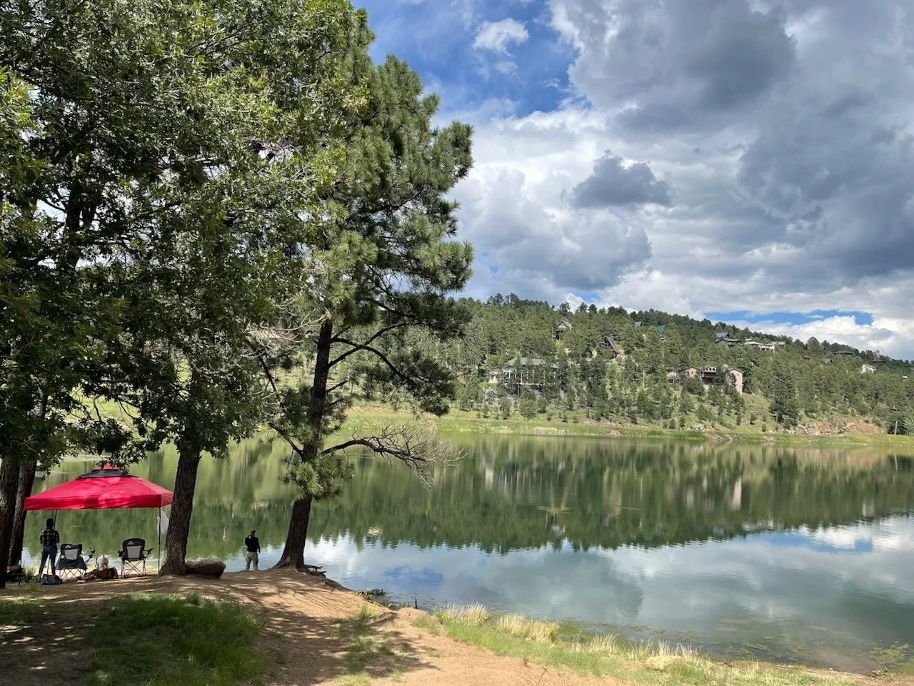 Visitors enjoying a shaded picnic spot beside Alto Lake, with still water, forested hills, and cabins in the distance