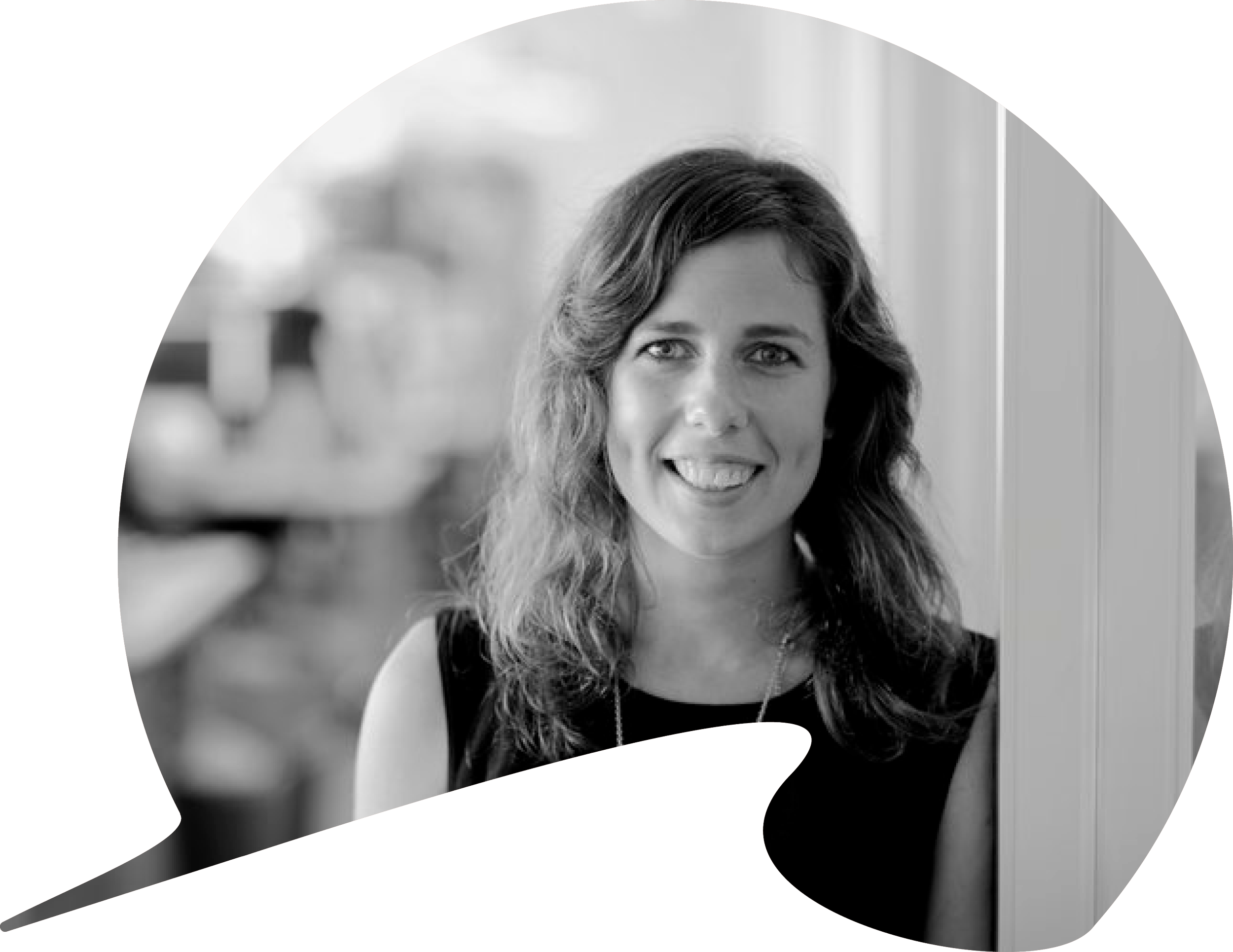 Black and white photo of a woman with wavy hair smiling, standing near a window or door frame, with a blurred indoor background.