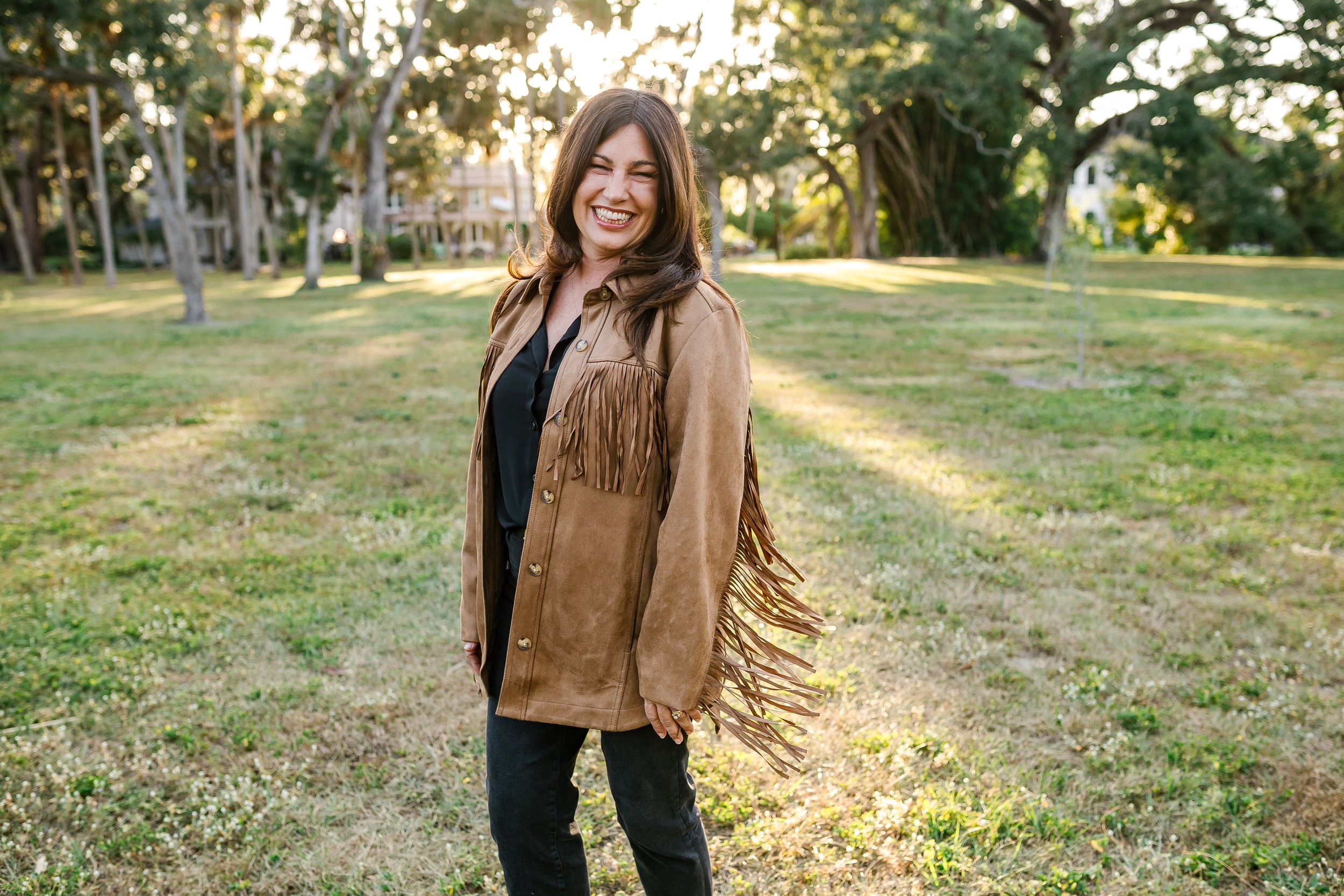 A woman with brown hair smiling and squinting, wearing a tan fringed jacket and black shirt, standing outdoors in a park with green grass and trees in the background, during sunset.