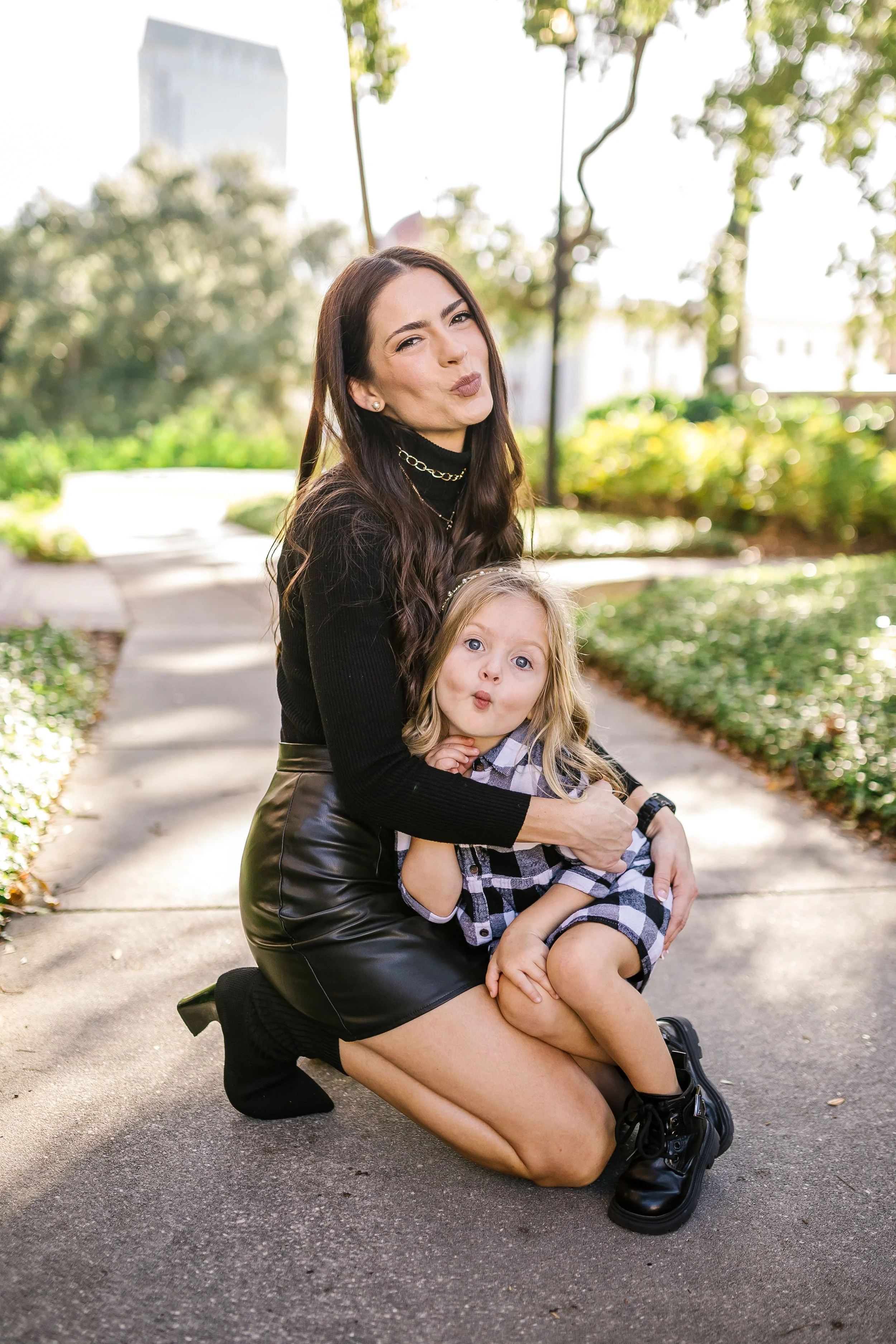 A woman with long dark hair and a black leather skirt kneels on a sidewalk, holding a young girl with blonde hair and blue eyes. The woman makes a playful kissy face while the girl looks surprised and curious. They are outdoors in a park with green t