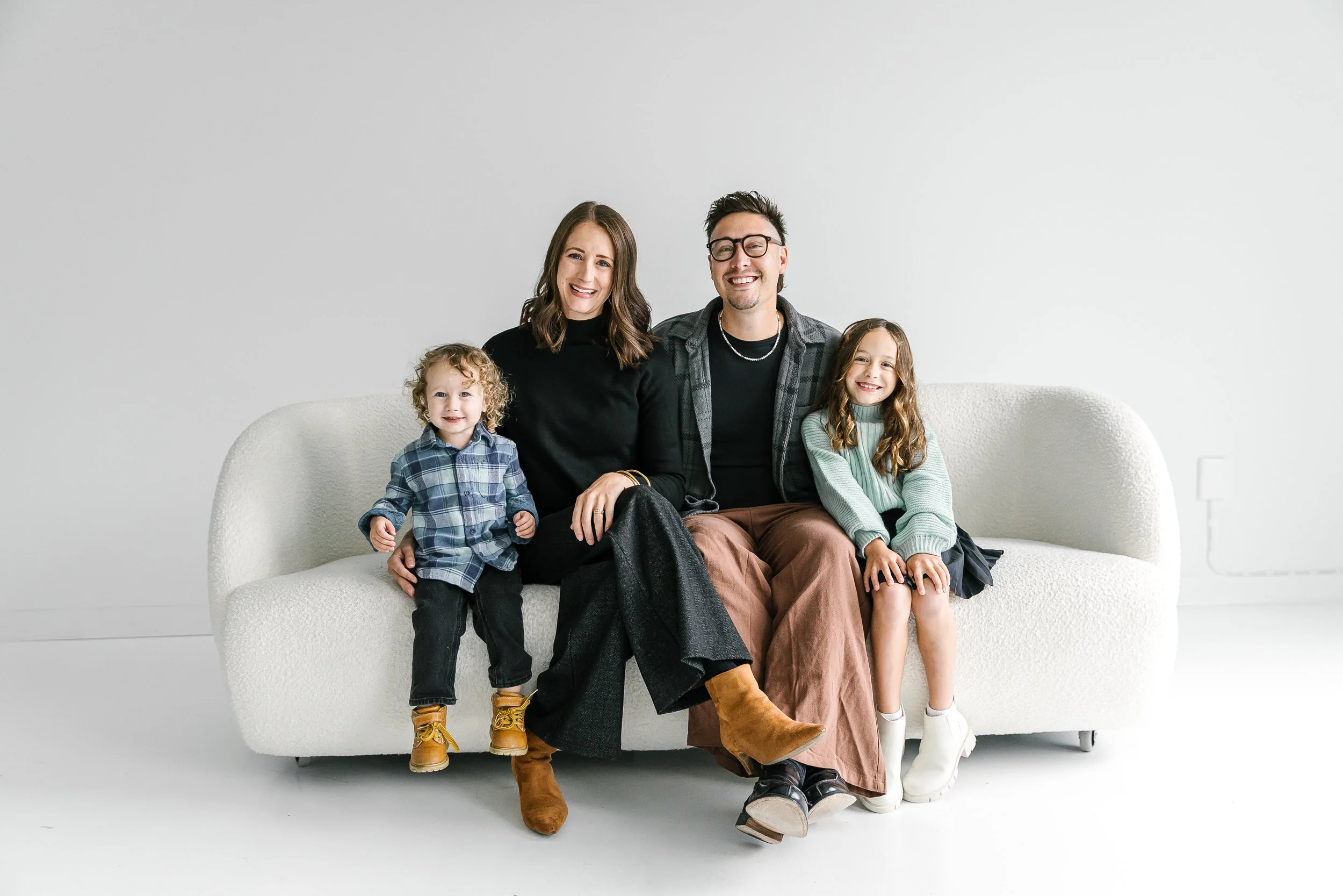 Family sitting on a white couch in a minimalistic room, smiling at the camera. The family includes two adults and two children, all dressed casually.