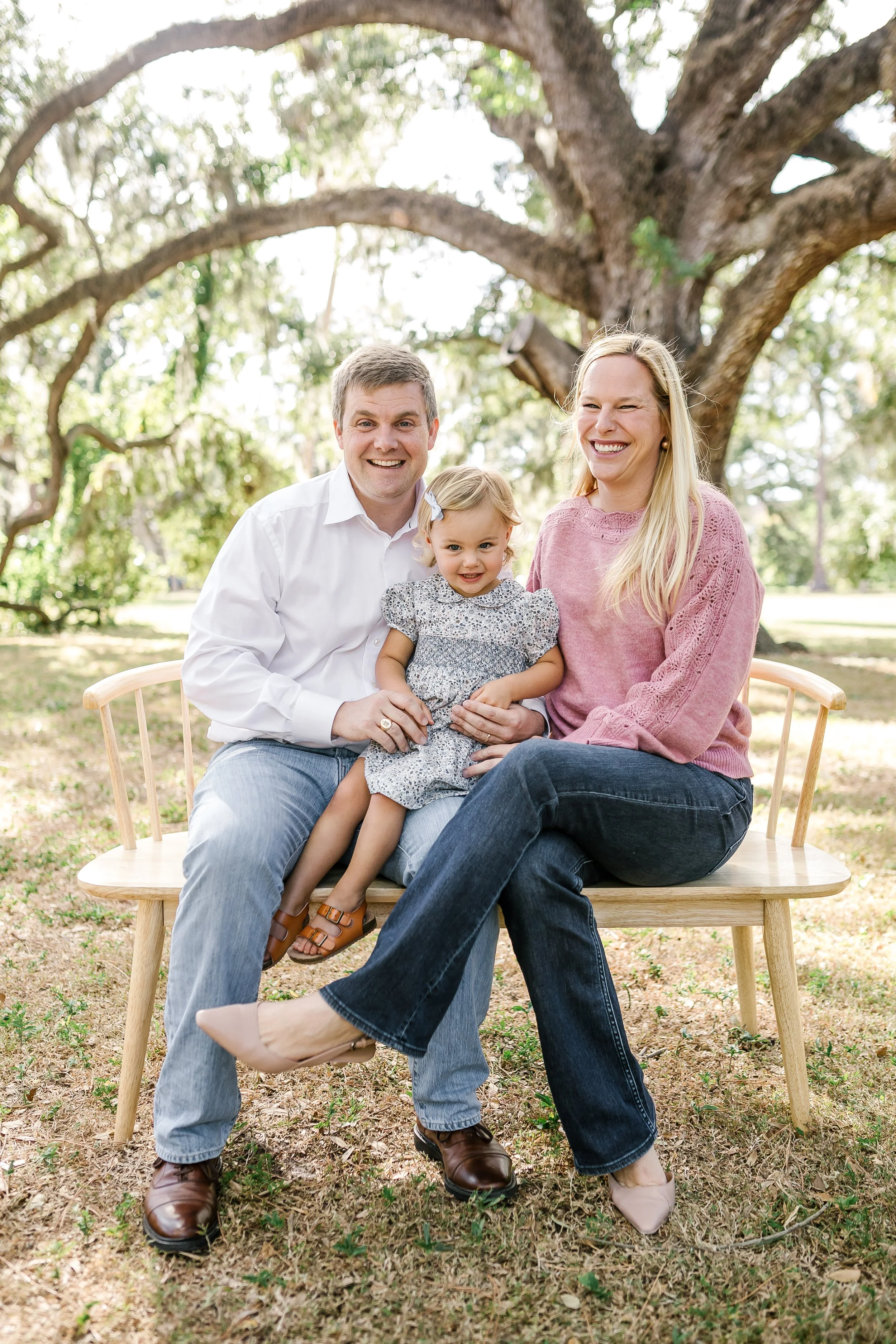 A family of three sitting on a wooden bench outdoors under a large tree, smiling and enjoying a sunny day.