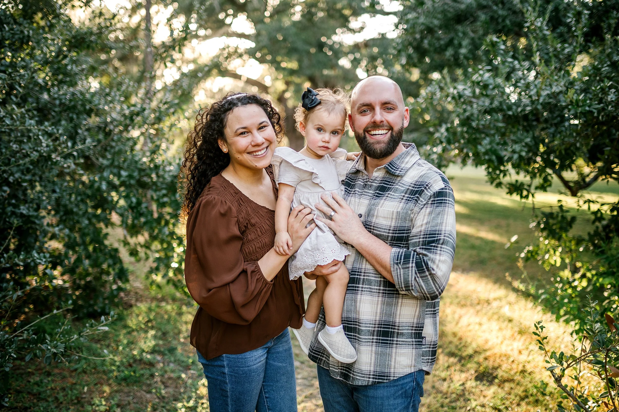 A happy family of three, a woman, a man, and a young girl, standing outdoors in a green park with trees in the background, during daylight.