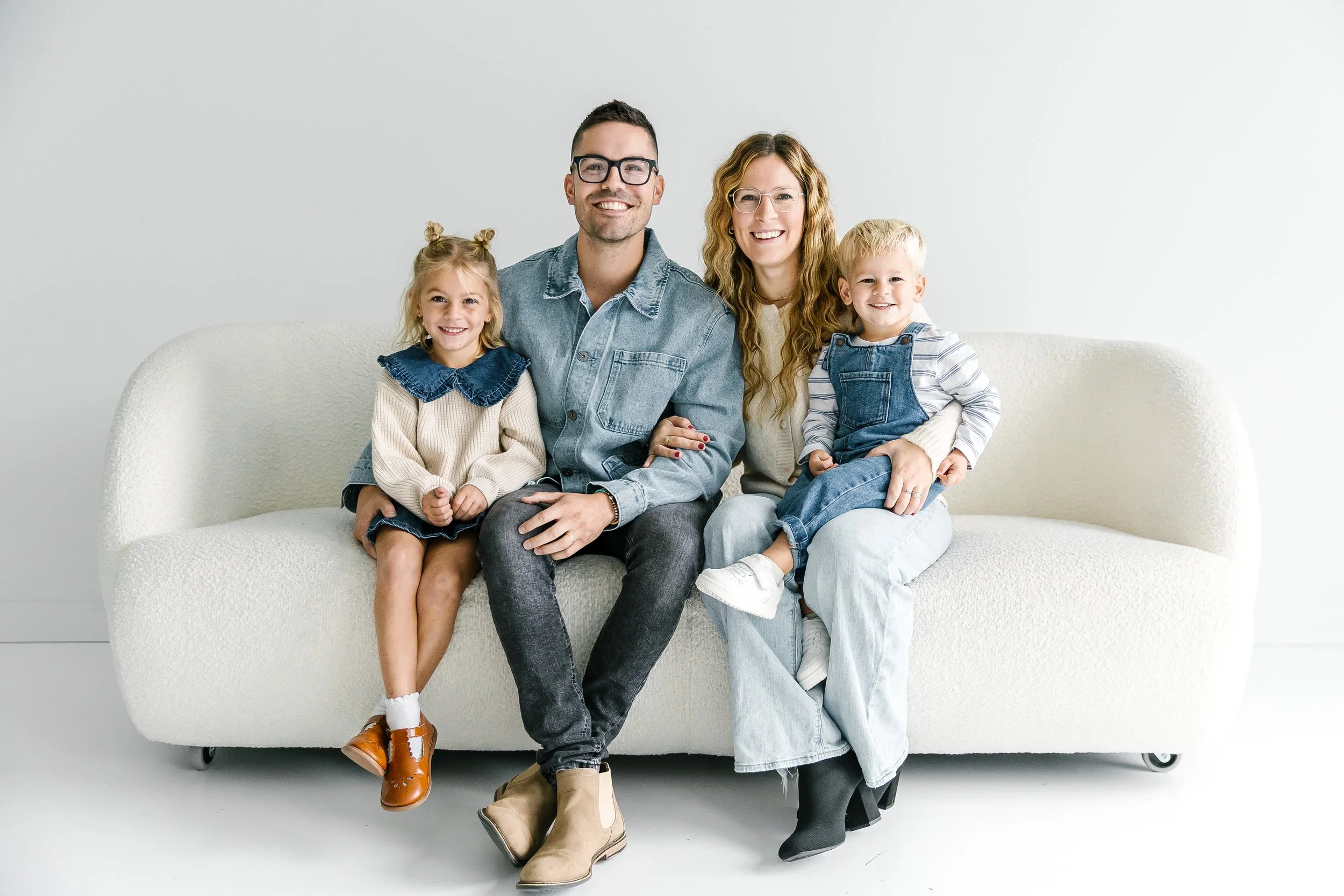 A family of four sitting on a white couch against a plain white wall, smiling for the camera. The father wears glasses and a denim shirt, the mother has long curly hair and glasses, a young girl with two buns in her hair sits on the father's lap, and a young boy with short blond hair is sitting on the mother's lap, wearing denim overalls.