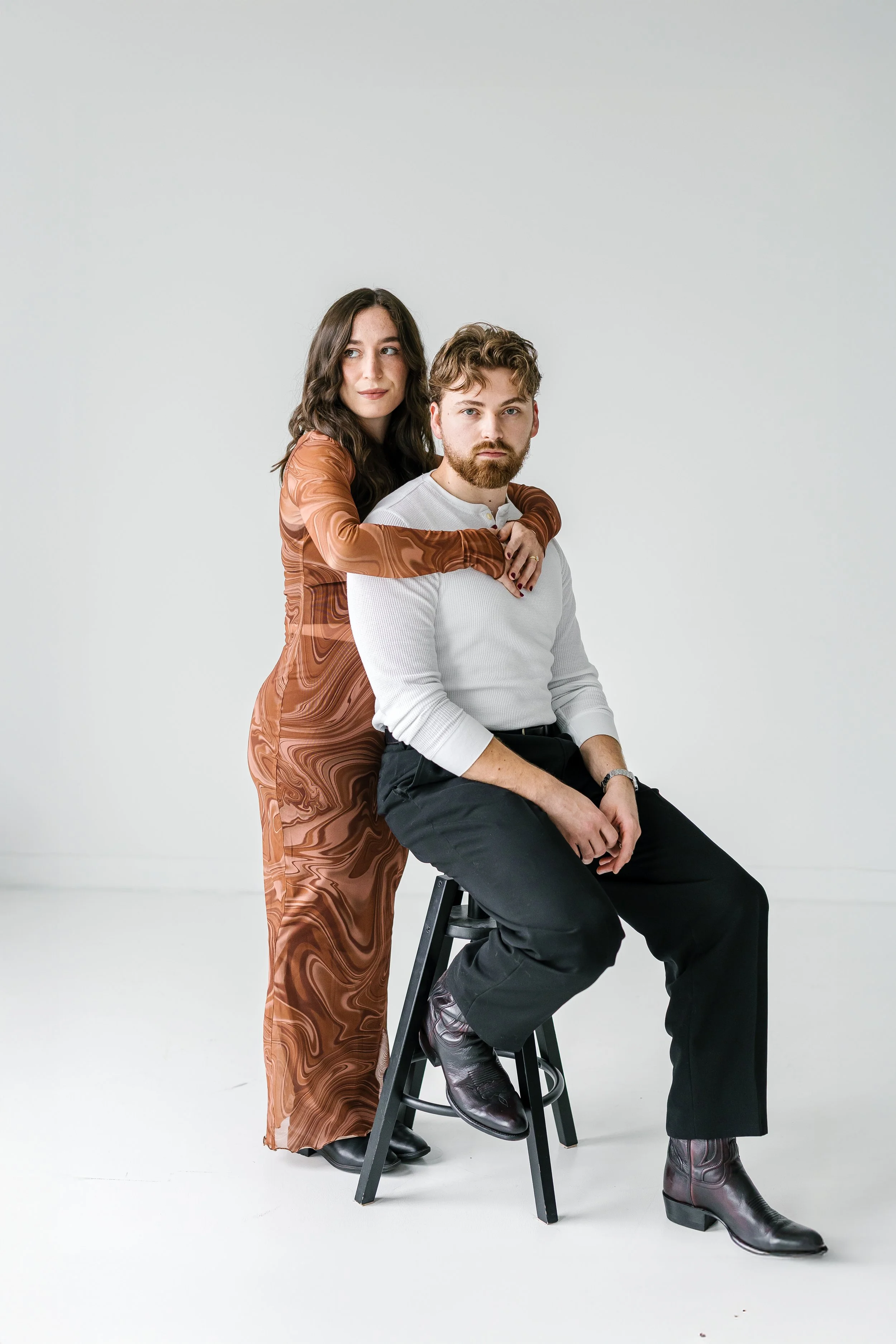 A man with a beard and a woman with long dark hair posing together in a studio with a light gray background. The woman stands behind the man, resting her arms on his shoulders, while he is seated on a black stool, wearing black pants and boots.