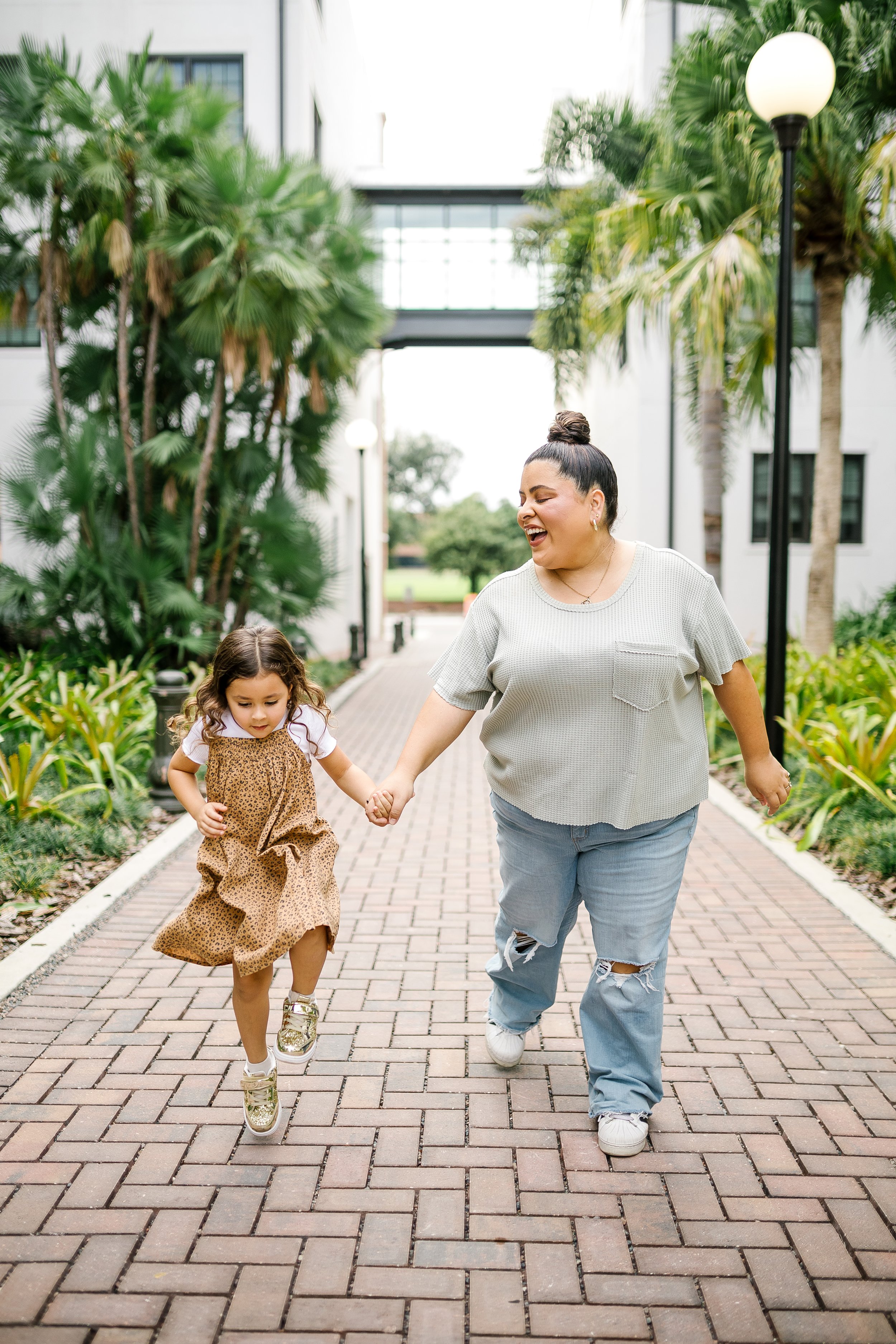 A woman and a young girl are walking hand in hand on a brick pathway in an outdoor setting with lush green trees and plants. The woman is laughing and wearing ripped jeans and a gray t-shirt, while the girl is wearing a leopard print dress and gold s