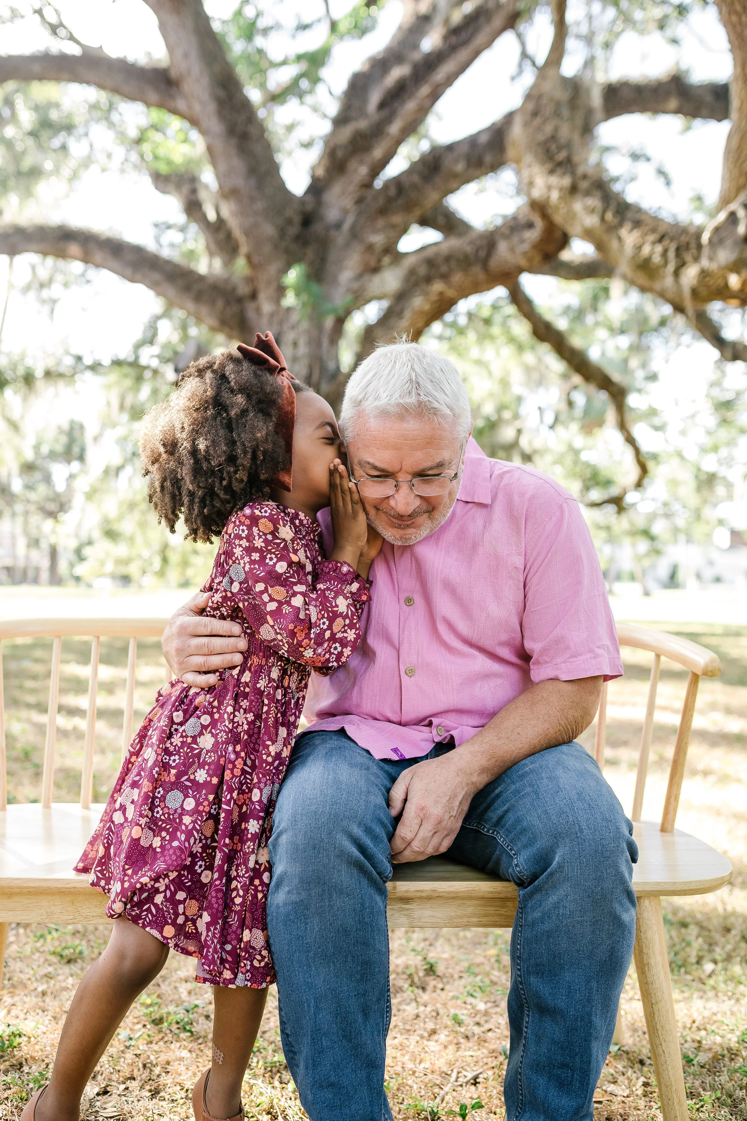 A young girl whispering into an older man's ear while sitting on a wooden bench outdoors under a large tree.