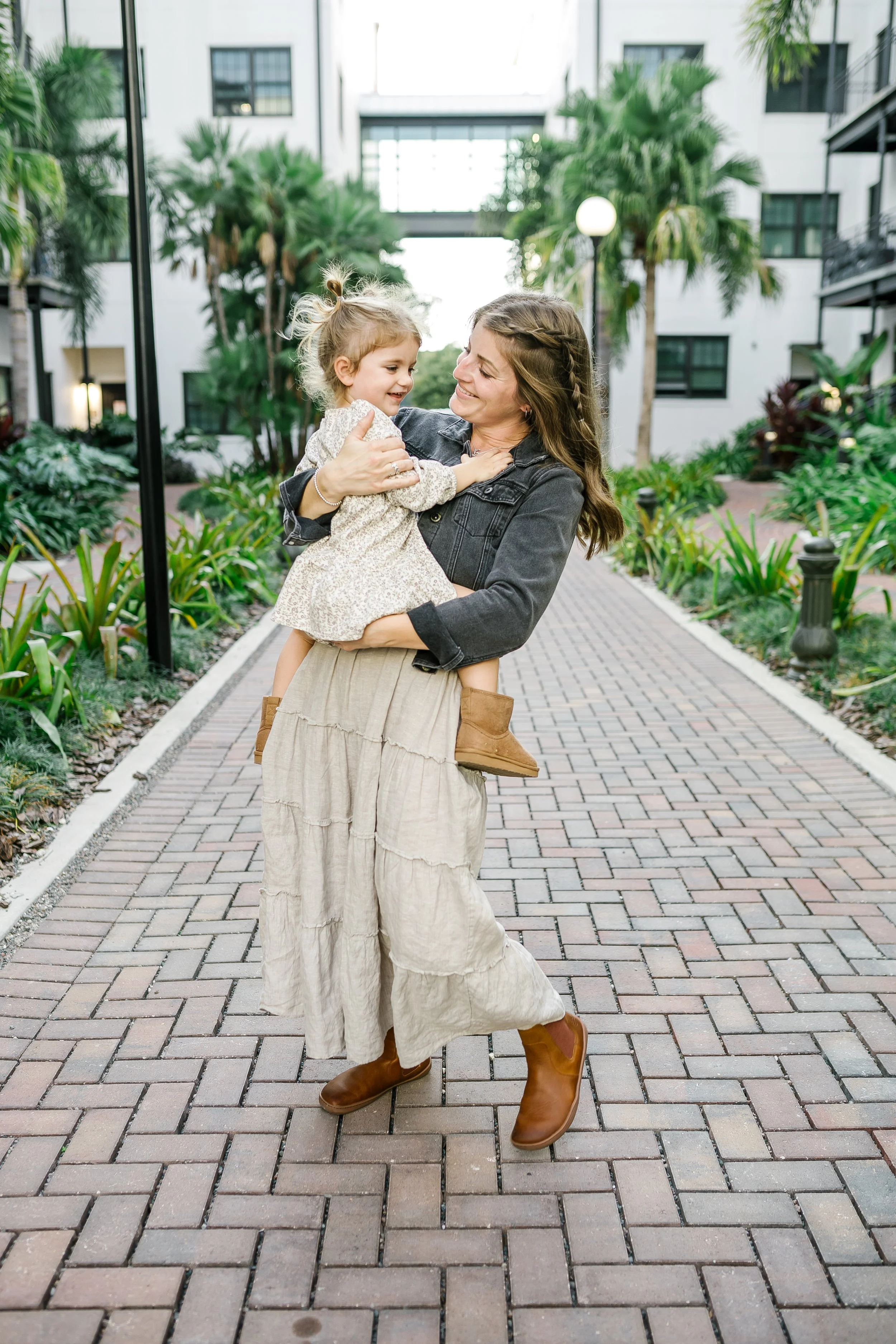 A woman in a long beige skirt and brown boots holding a smiling young girl in a light-colored dress, walking in a courtyard with plants and white buildings in the background.