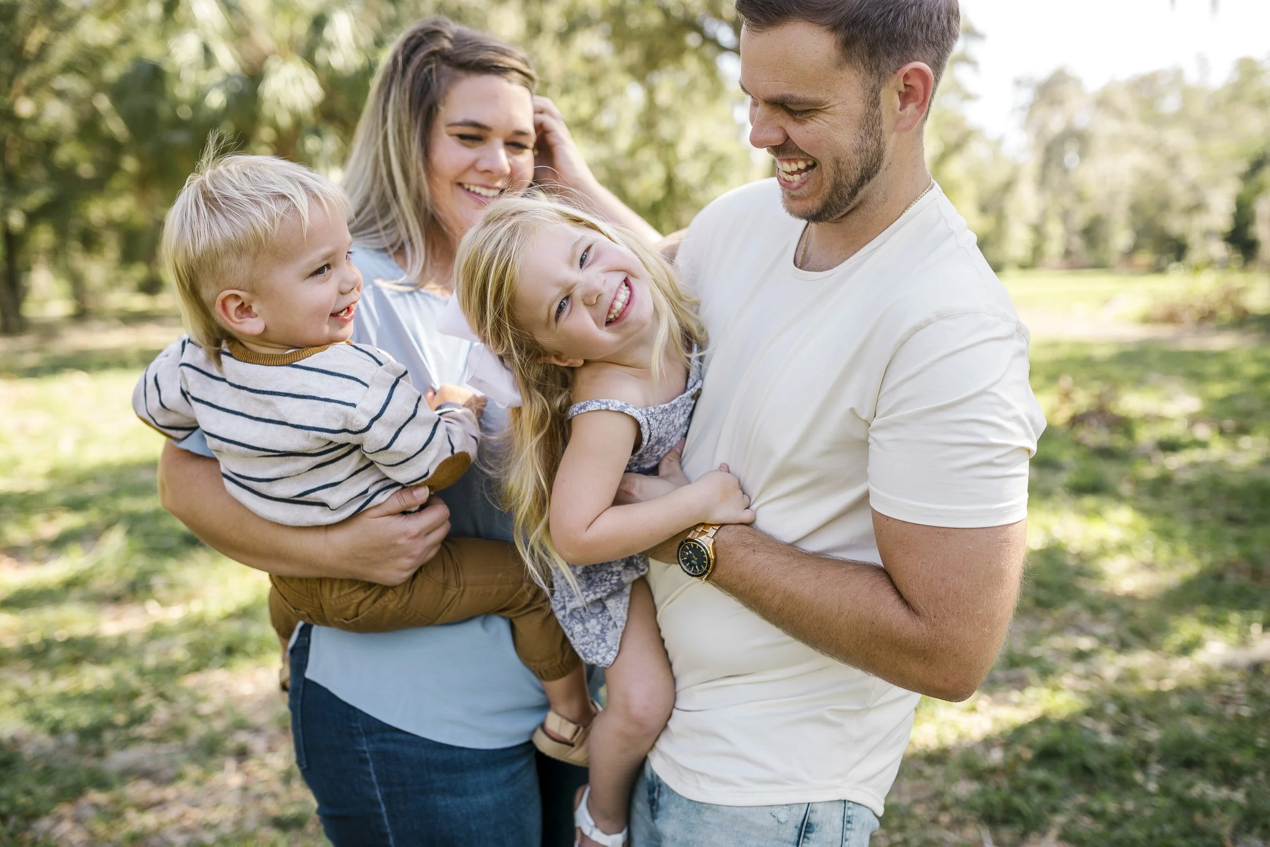 Family of four with two young children, a boy and a girl, enjoying an outdoor moment together in a park on a sunny day, all smiling and laughing.