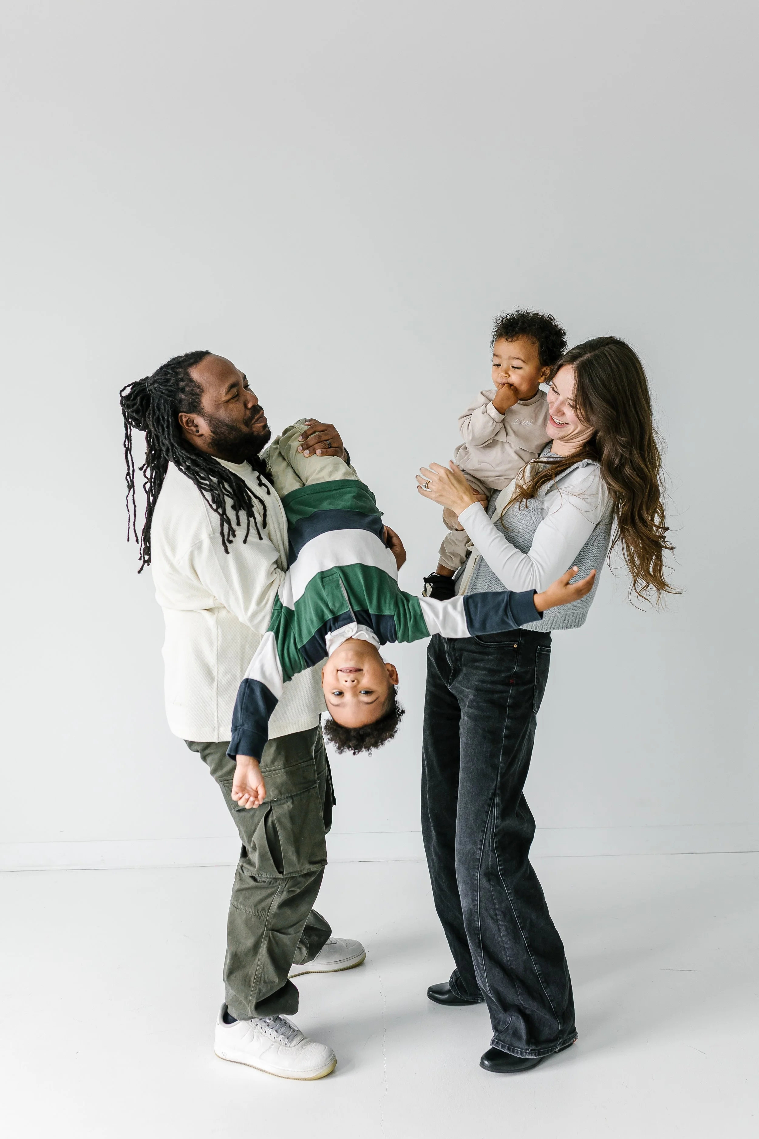 A family of four, including a man with long dreadlocks, a woman with long wavy hair, and two young children, one boy hanging upside down and a girl being held by the woman, all smiling and interacting in a minimal white room.