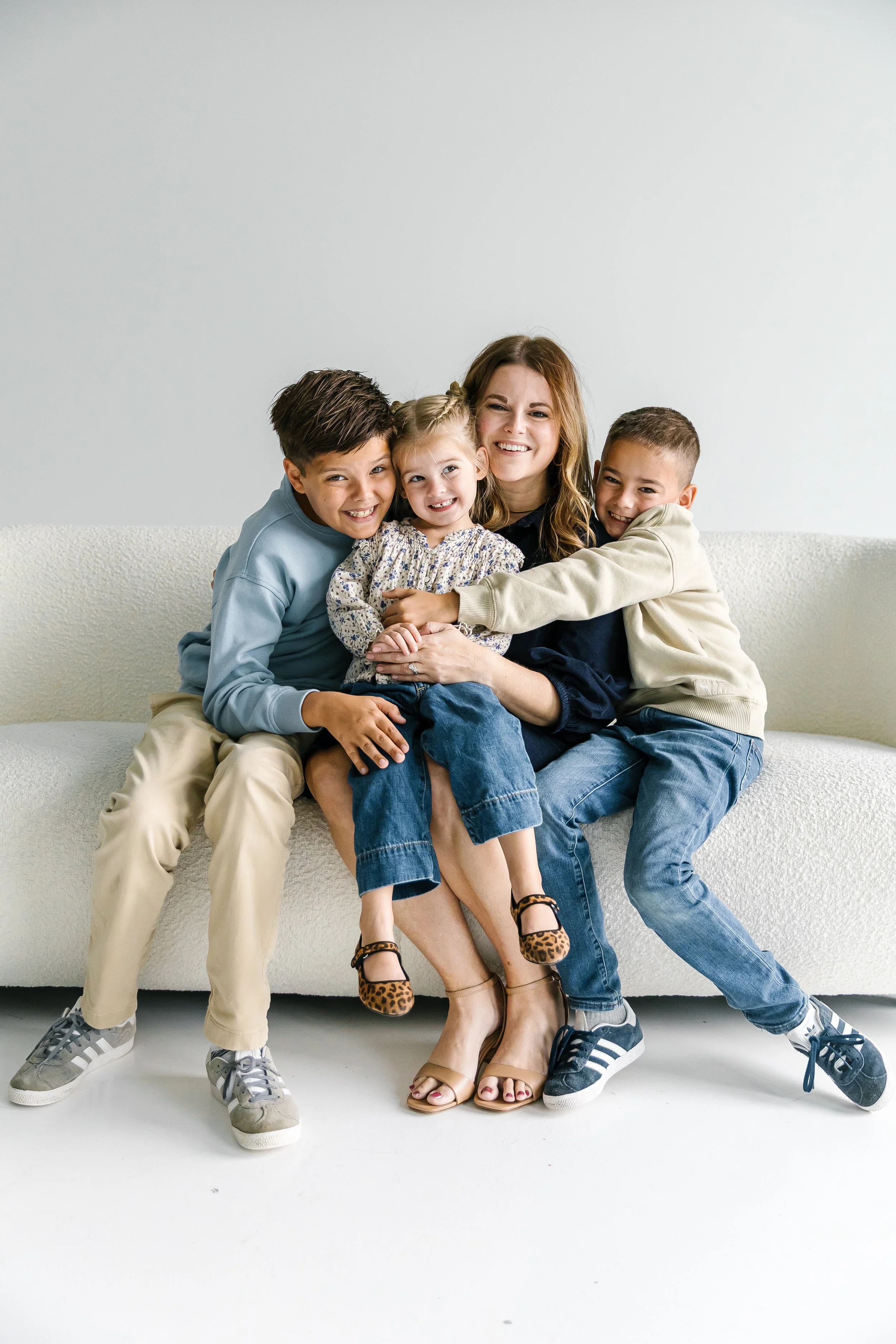 A woman and three children sitting on a white couch, smiling and hugging each other.