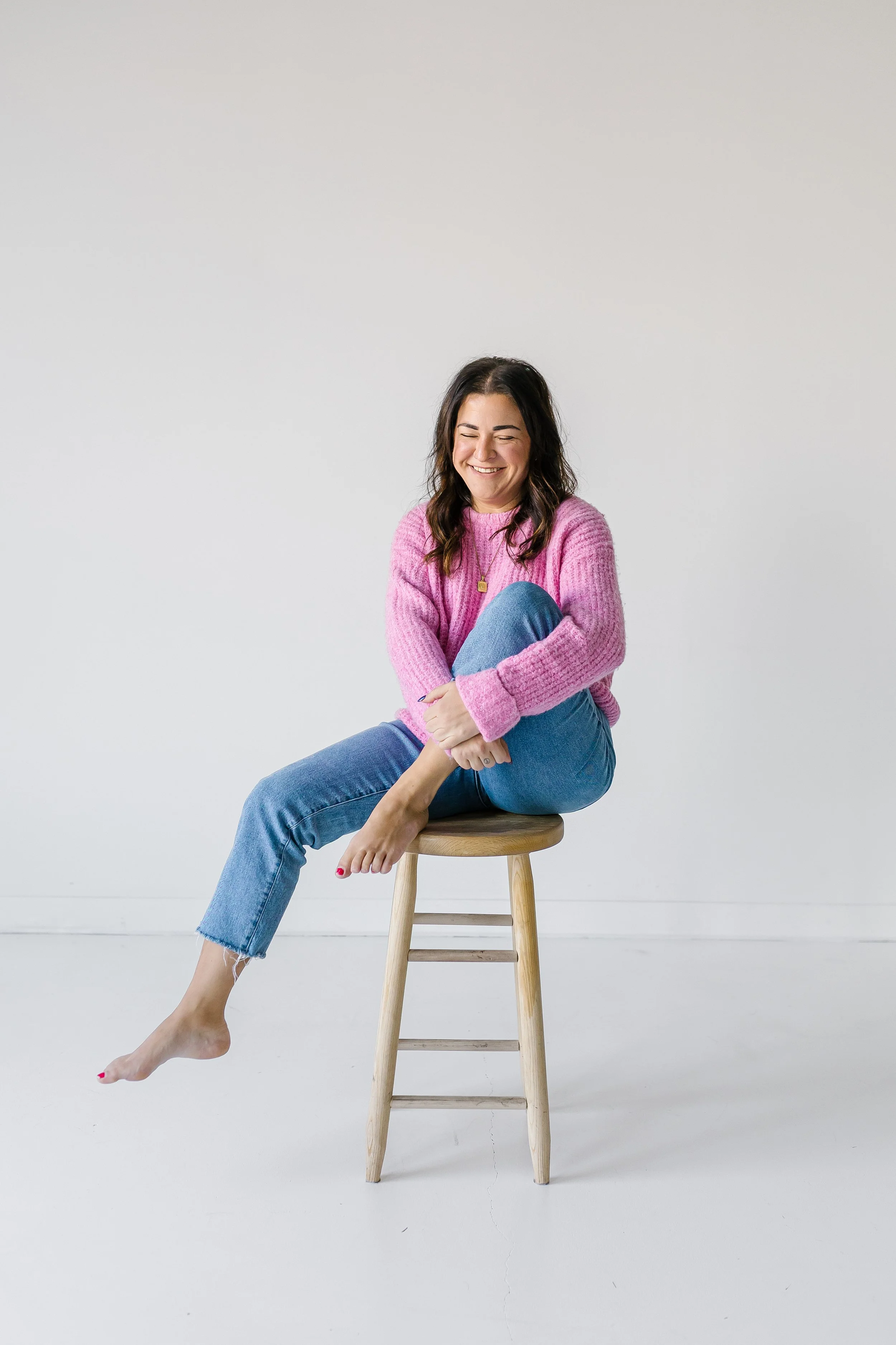 A woman with dark hair, wearing a pink sweater and blue jeans, sitting on a wooden stool with crossed legs and smiling with her eyes closed against a plain white background.