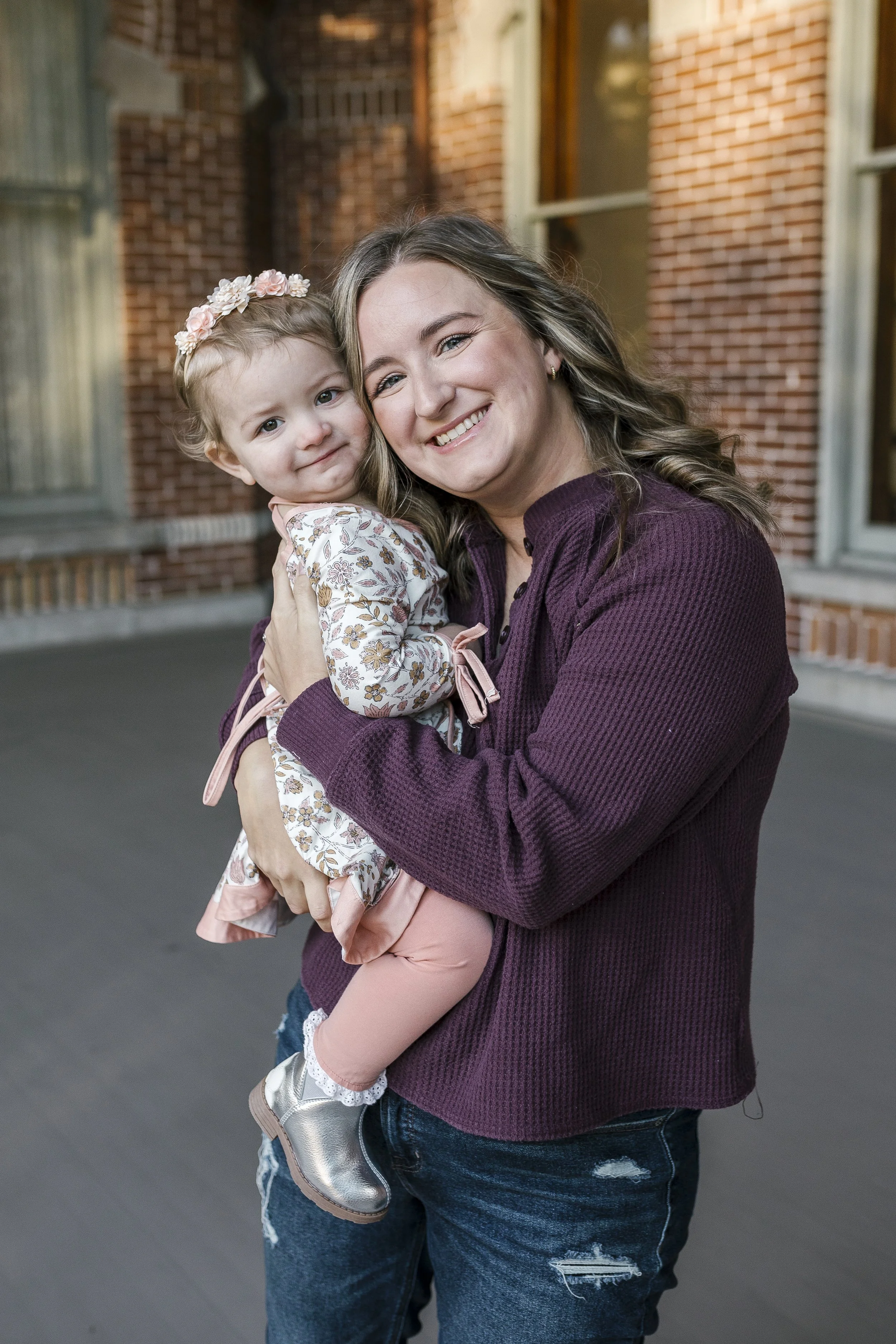 A woman with wavy blonde hair smiling and holding a young girl with curly blonde hair and a floral headband. They are outdoors in front of a red brick building.