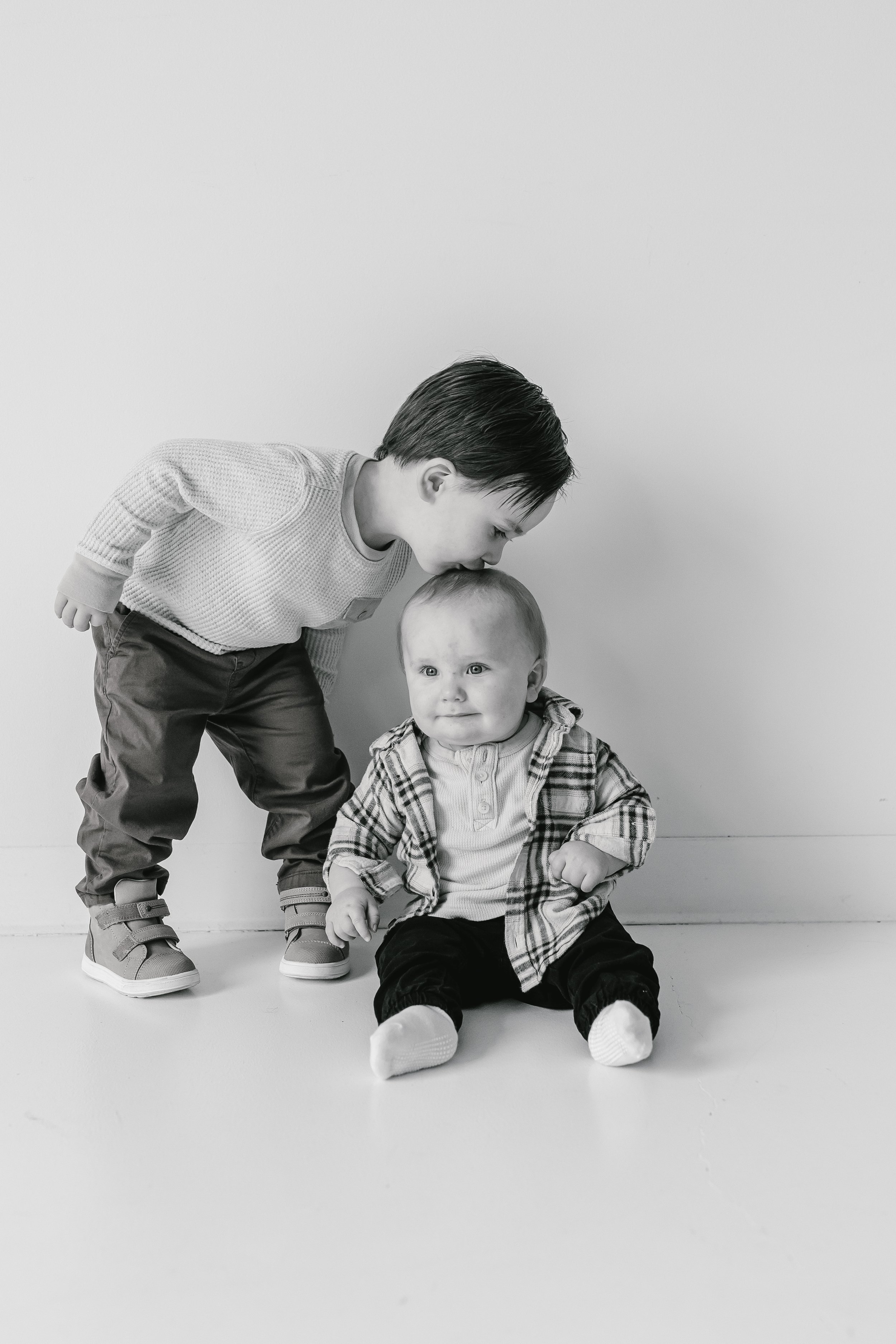 Two young boys, one standing and leaning over, the other sitting on the floor, both with short dark hair, in front of a plain white wall.