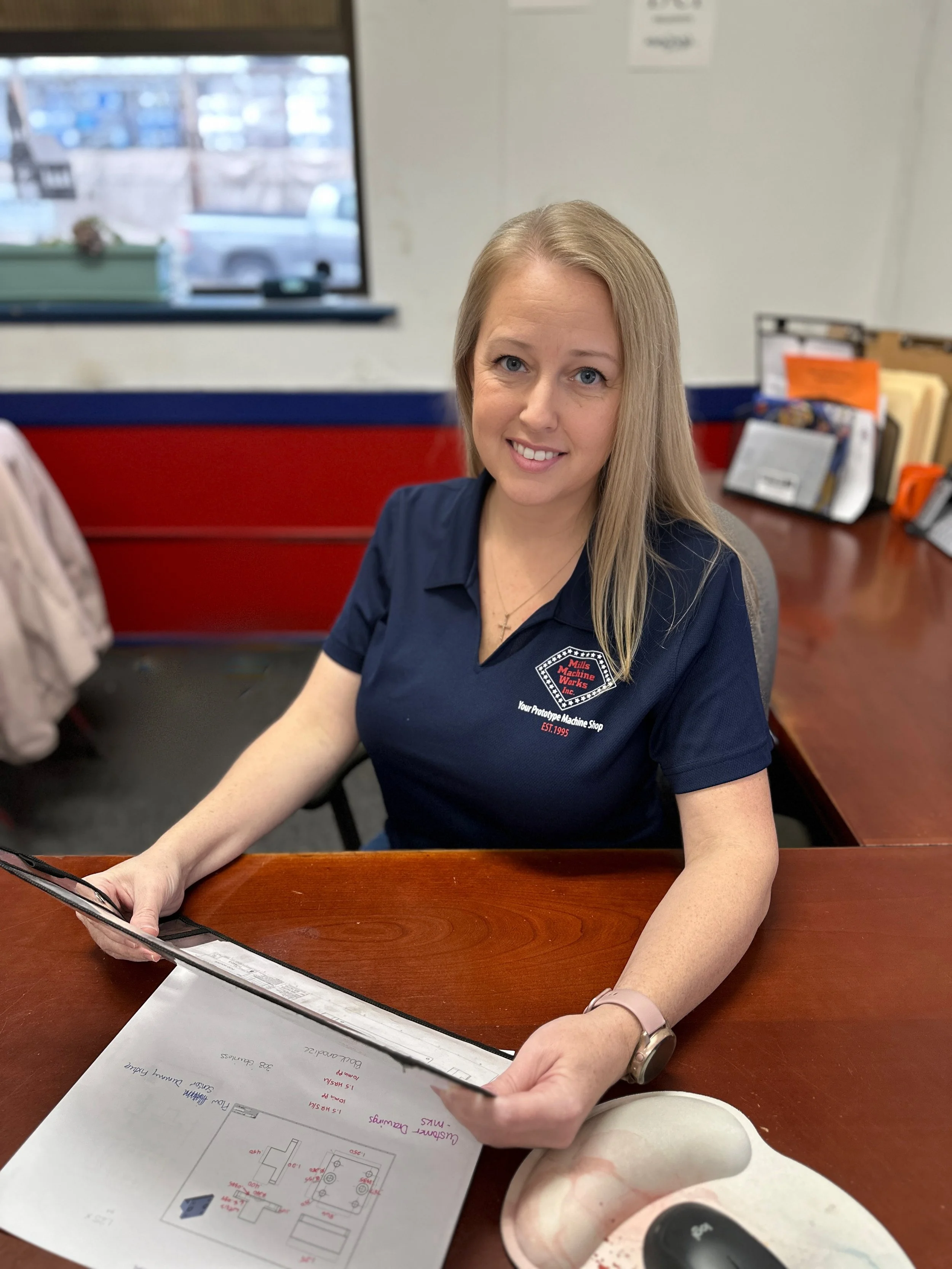 A woman sitting at a desk in an office setting, holding a document with diagrams and notes. She is wearing a navy blue polo shirt with a logo on it. The desk has a mouse pad, computer mouse, and a stack of papers. A window and files are visible in the background.