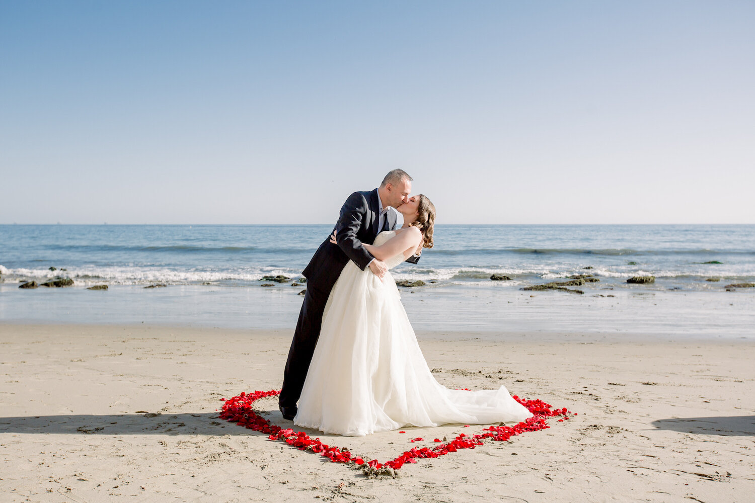 bride and groom kiss standing in a heart shape of floral petals on beach