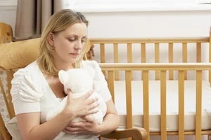 Blond woman in a nursery holding a white teddy bear, looking sad.