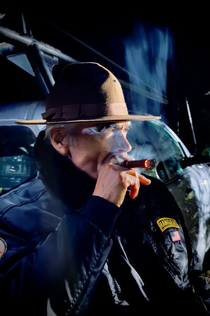 A woman wearing a ranger uniform and a brown wide-brimmed hat is smoking a cigar outdoors near a helicopter.