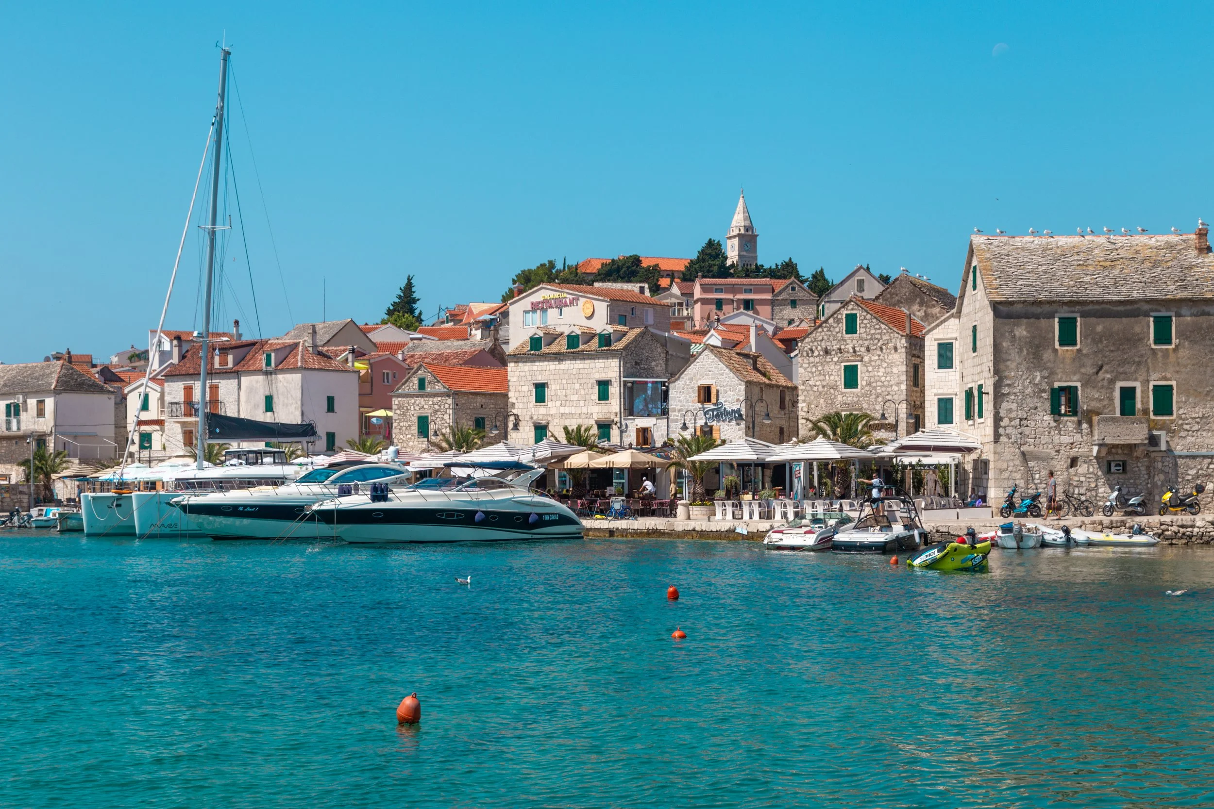 Harbor view with sailboats and motorboats docked in turquoise water, stone buildings with red-tiled roofs along the waterfront, outdoor cafes with umbrellas, and a hillside with more buildings and a church steeple in the background.