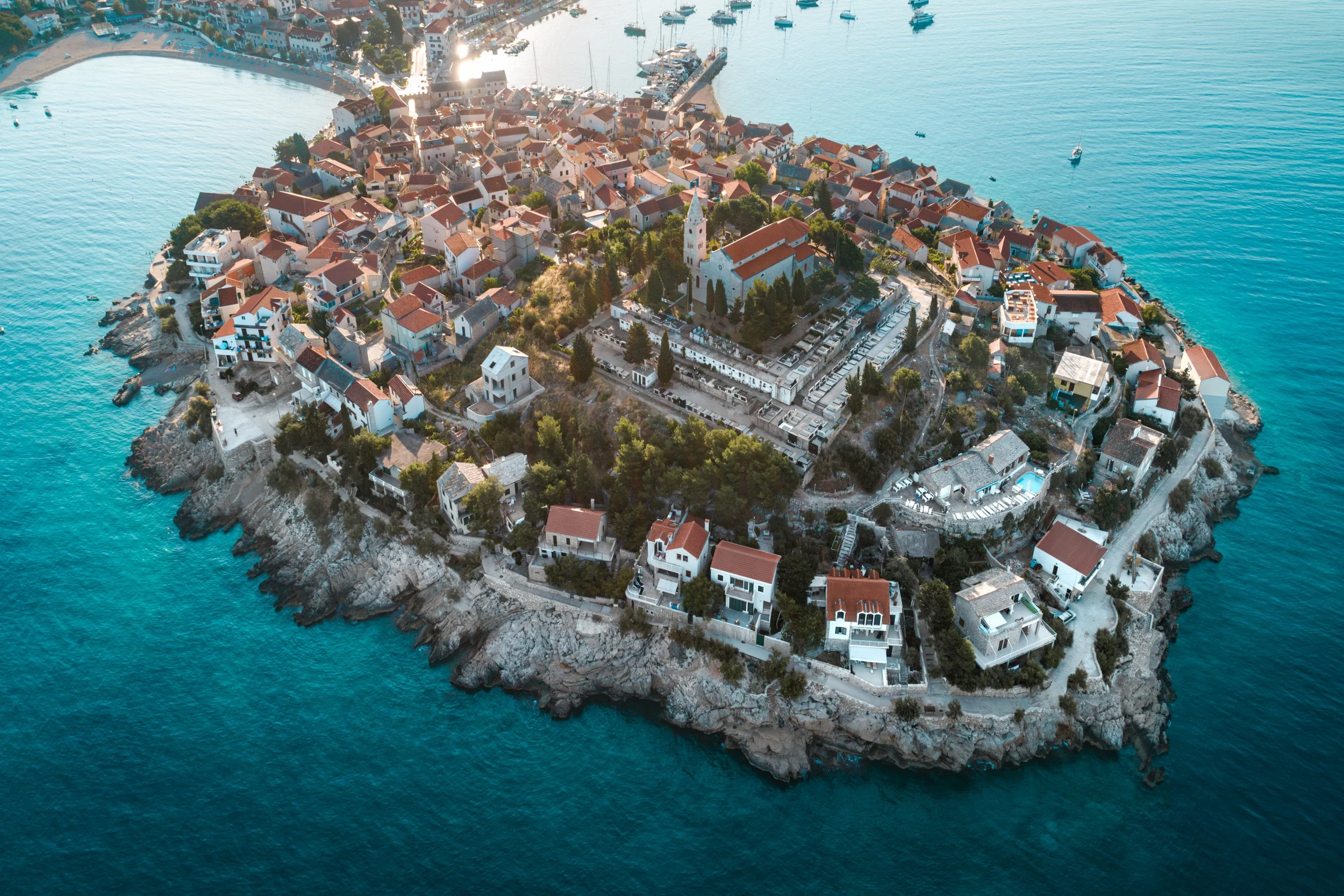 Aerial view of a small island town with red-roofed houses, a church, and various buildings surrounded by blue water.