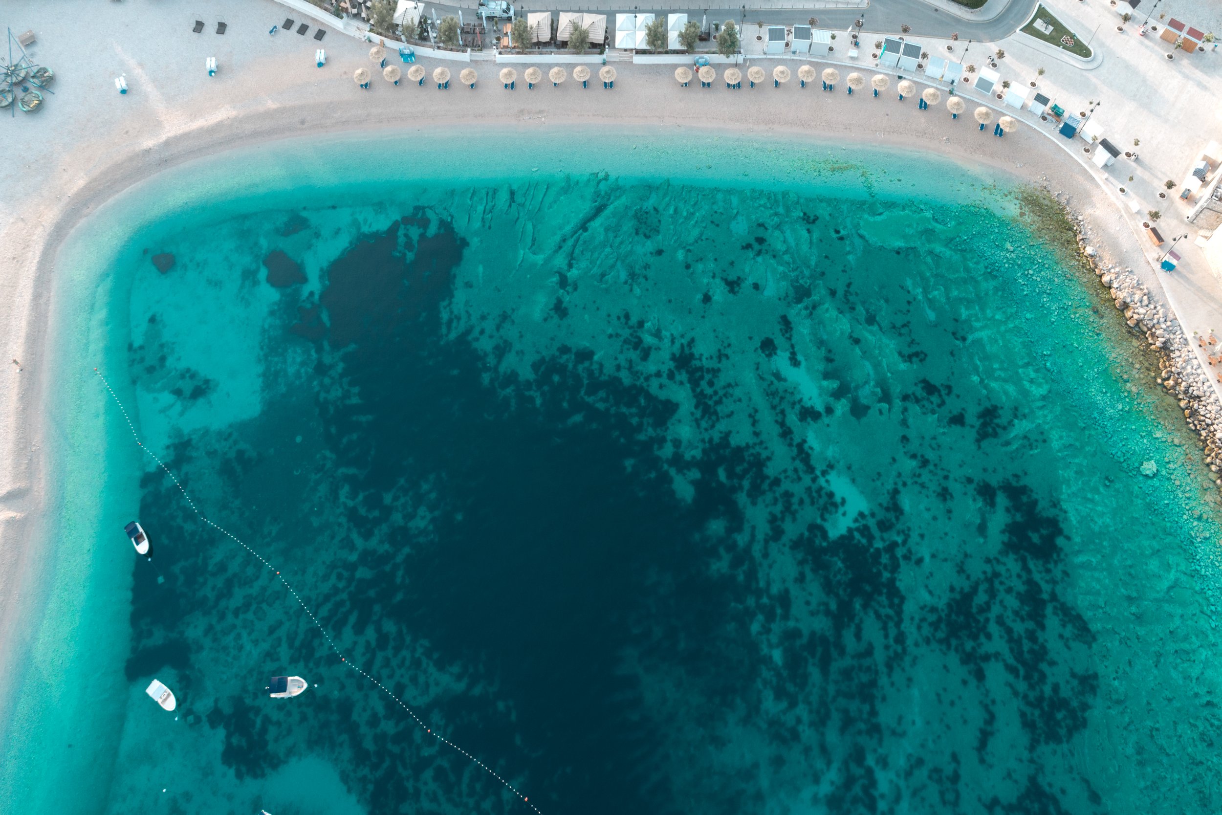 Aerial view of a beach with turquoise water, boats anchored near the shore, and a stone breakwater on the right. There are also beach umbrellas, sunbeds, and a paved walkway visible along the shoreline.