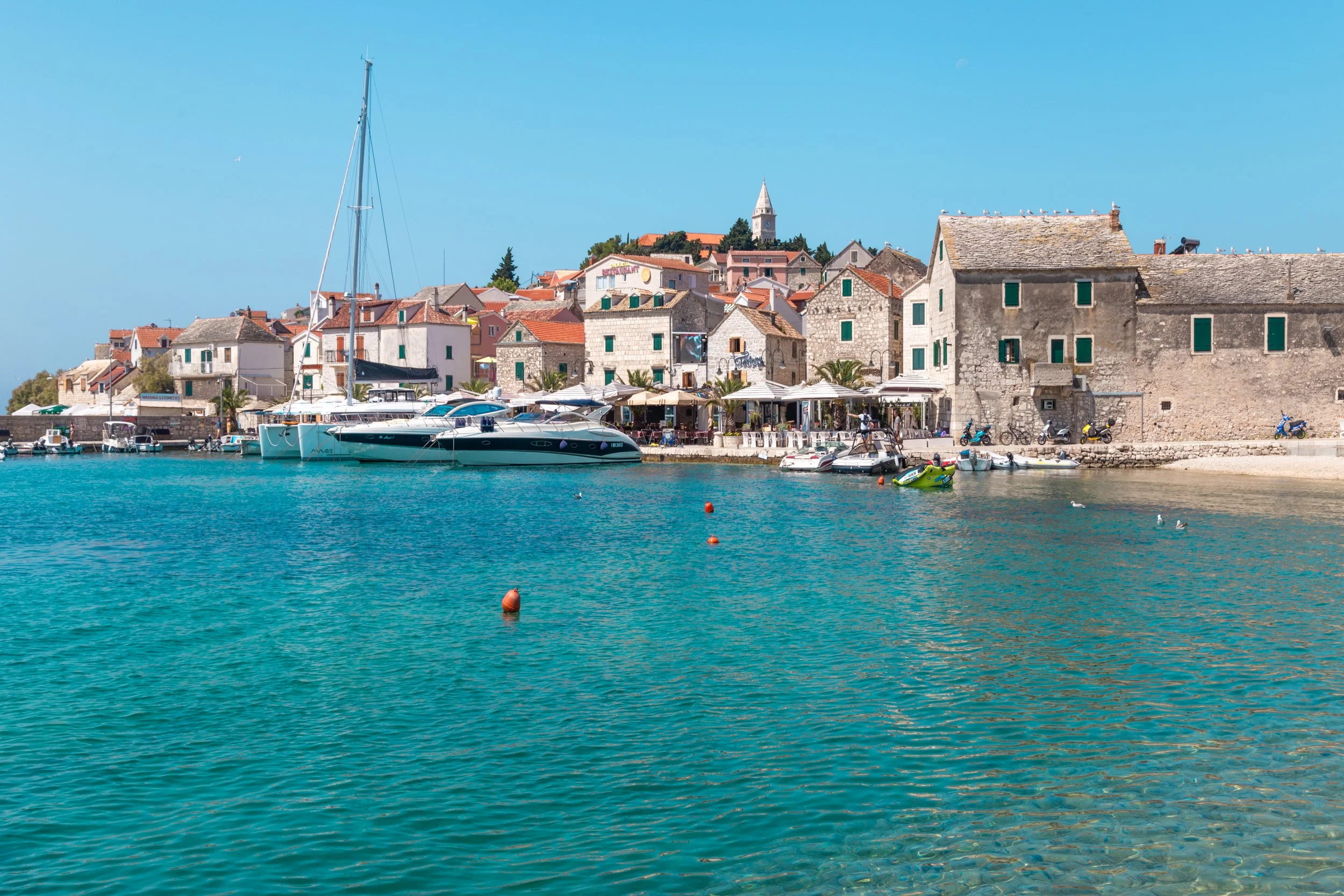 A picturesque coastal scene featuring a harbor with several boats, a row of seaside cafes and restaurants, and historic stone buildings with red-tiled roofs. Seagulls are visible in the sky and boats are docked along the waterfront.